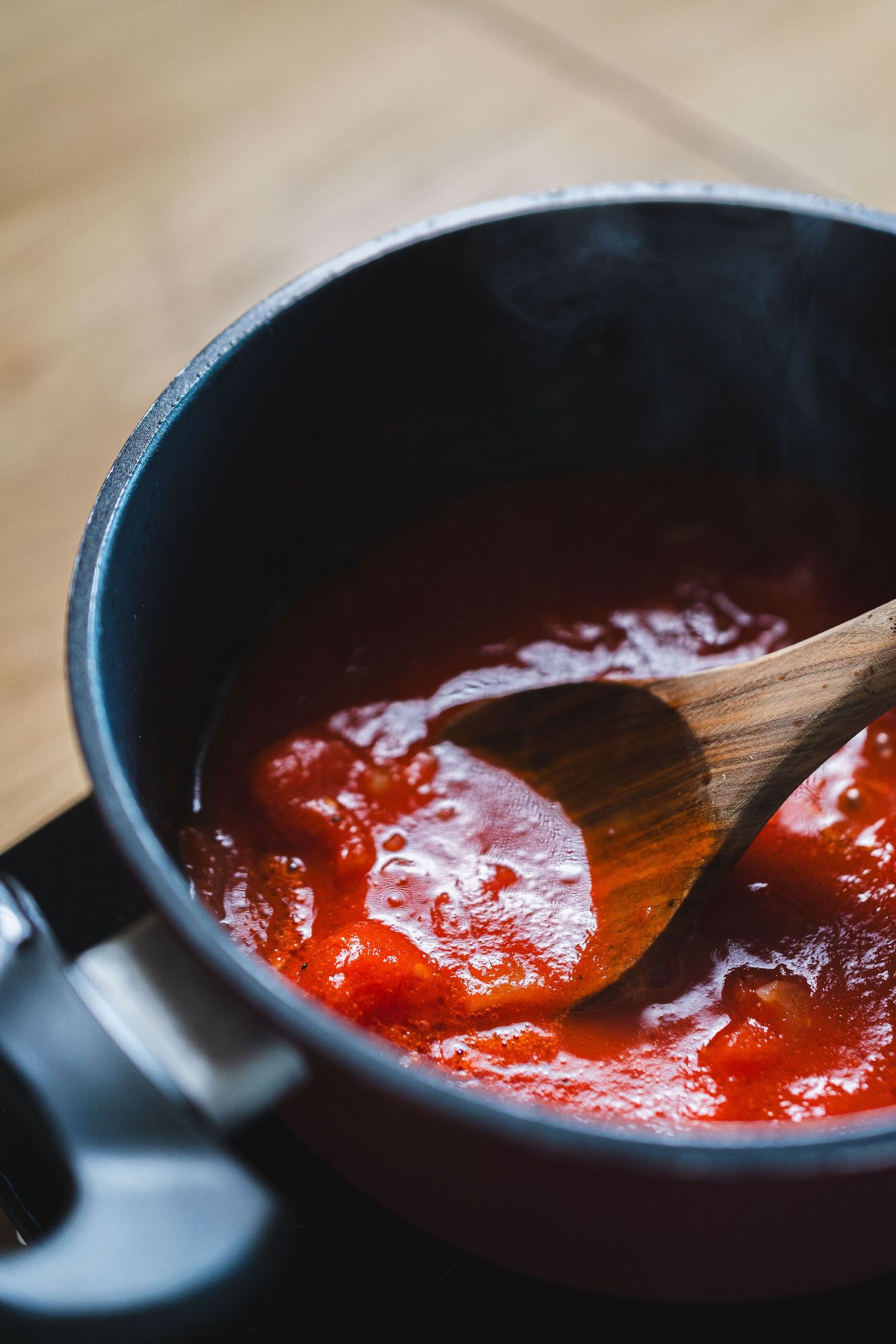 Tomato sauce simmering in dark saucepan being stirred with wooden spoon on stovetop.