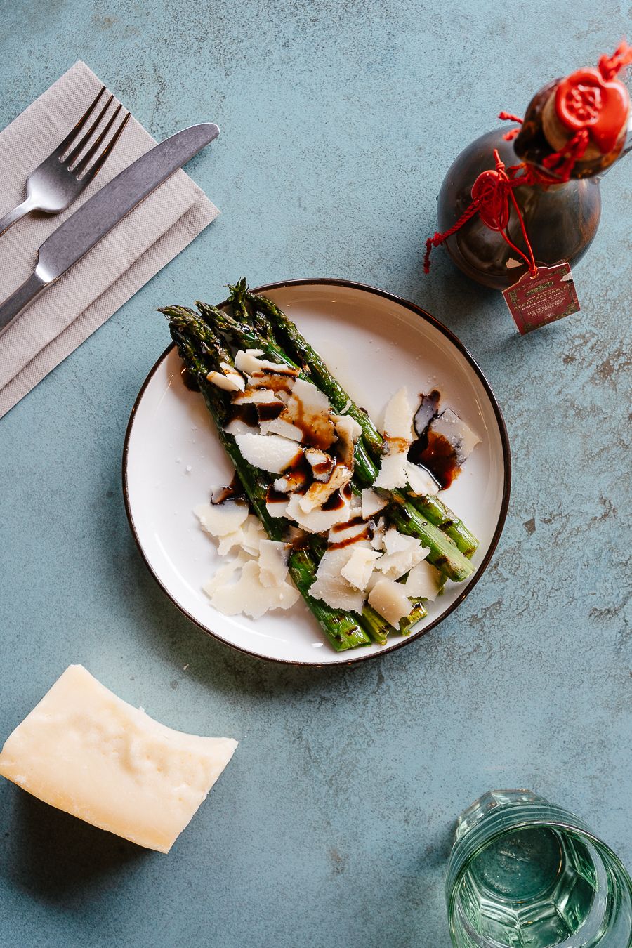 Grilled asparagus with parmesan shavings and balsamic glaze on plate with cutlery on blue surface.