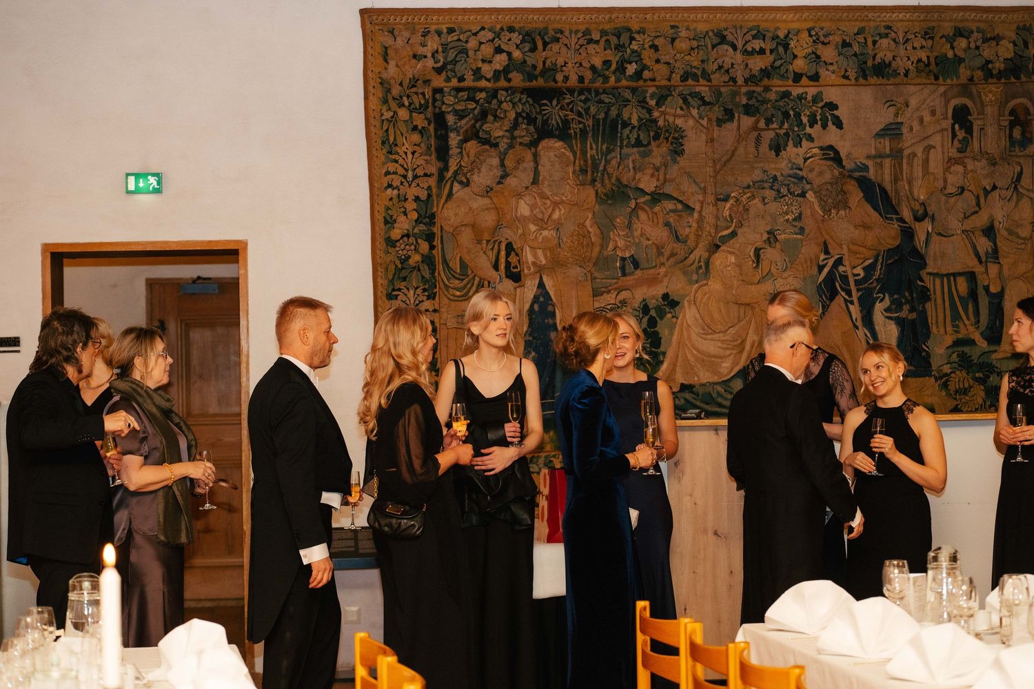 Elegantly dressed guests socializing near dining tables beneath historic tapestry at formal gala dinner.