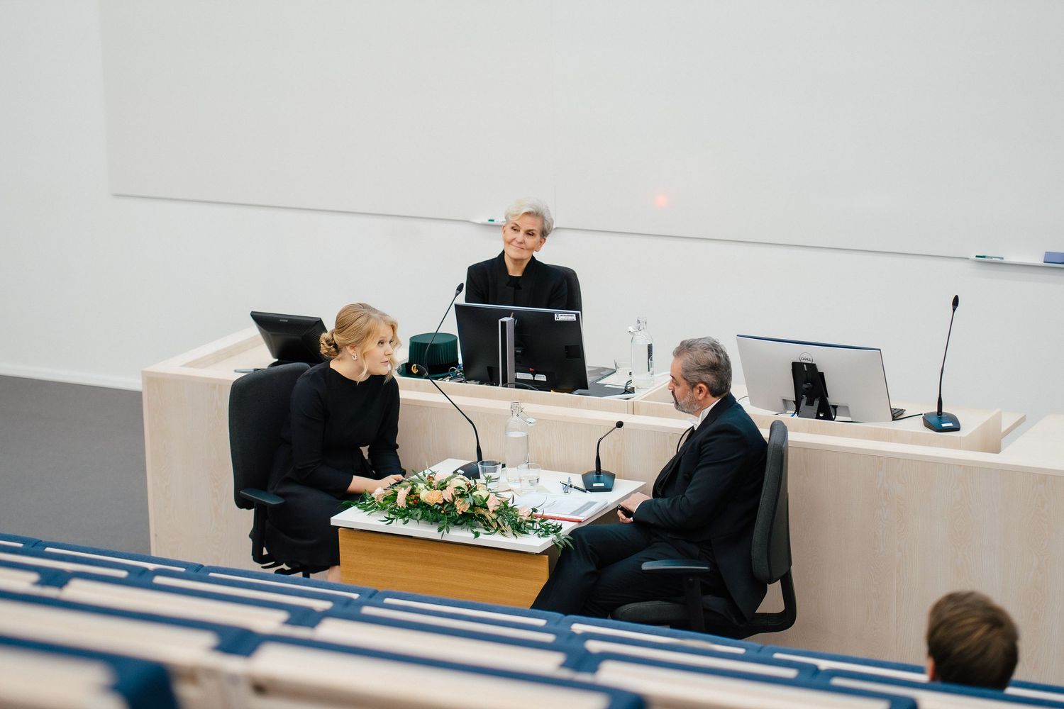 Three people in formal attire seated at decorated conference table with flowers during official meeting.