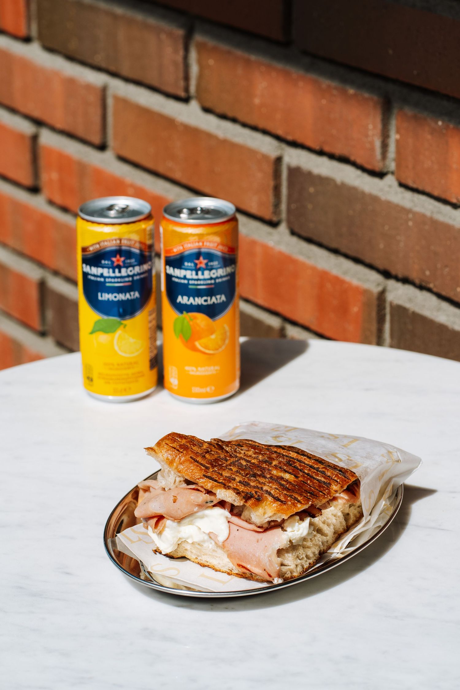 Grilled panini sandwich on plate with San Pellegrino Limonata and Aranciata cans on marble table.
