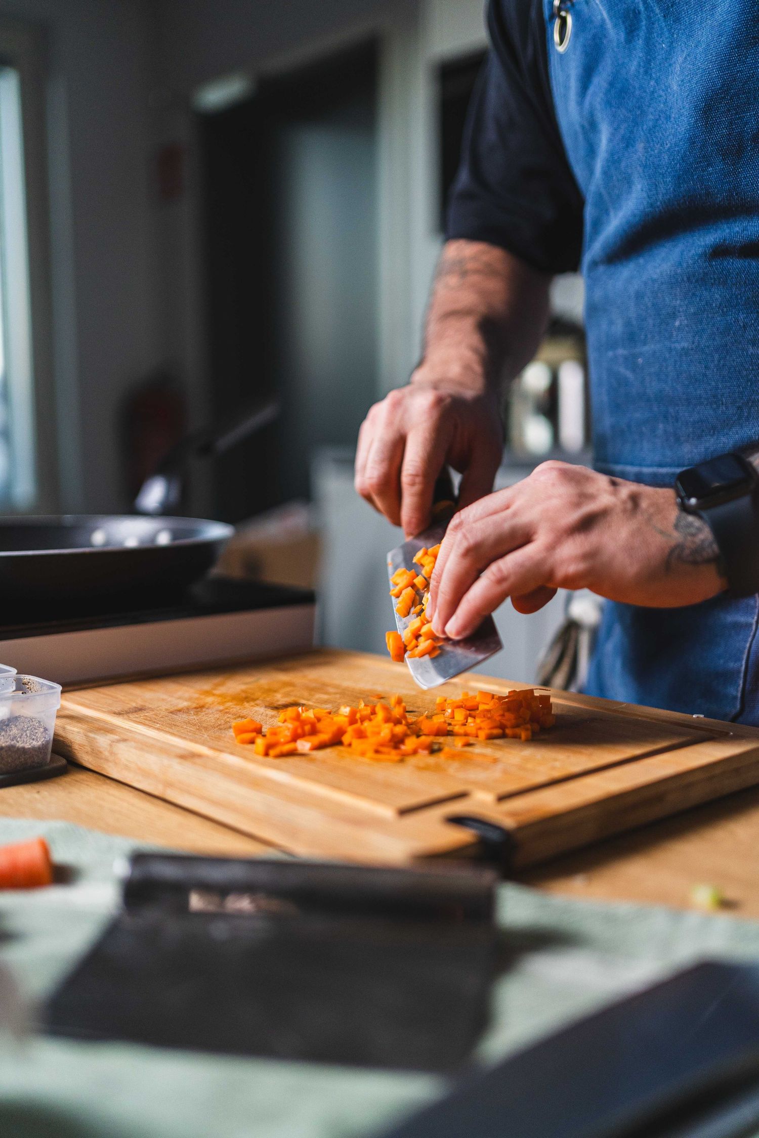 Chef in blue apron dicing carrots on wooden cutting board in professional kitchen setting.
