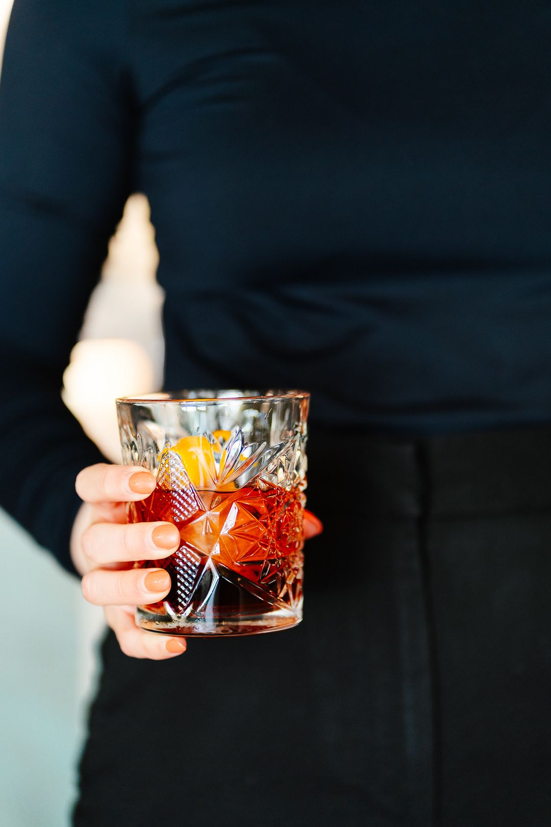 Person in black outfit holding a crystal old fashioned glass with dark cocktail and orange garnish.