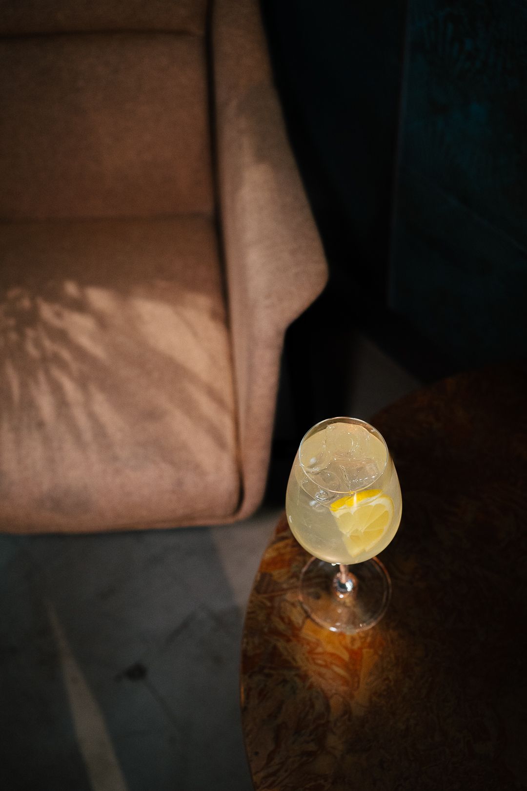 Elegant cocktail in stemmed glass with lemon garnish and ice on marble table beside brown sofa.