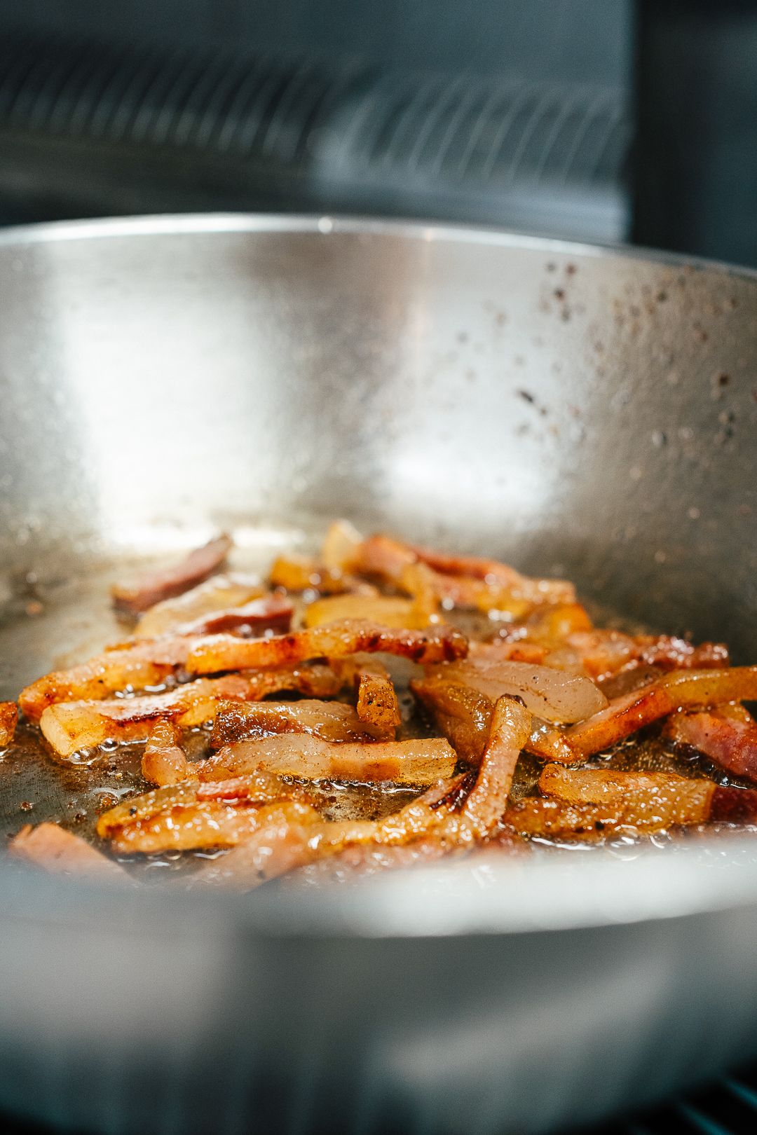 Crispy bacon strips sizzling in hot oil inside a stainless steel frying pan on a stove.