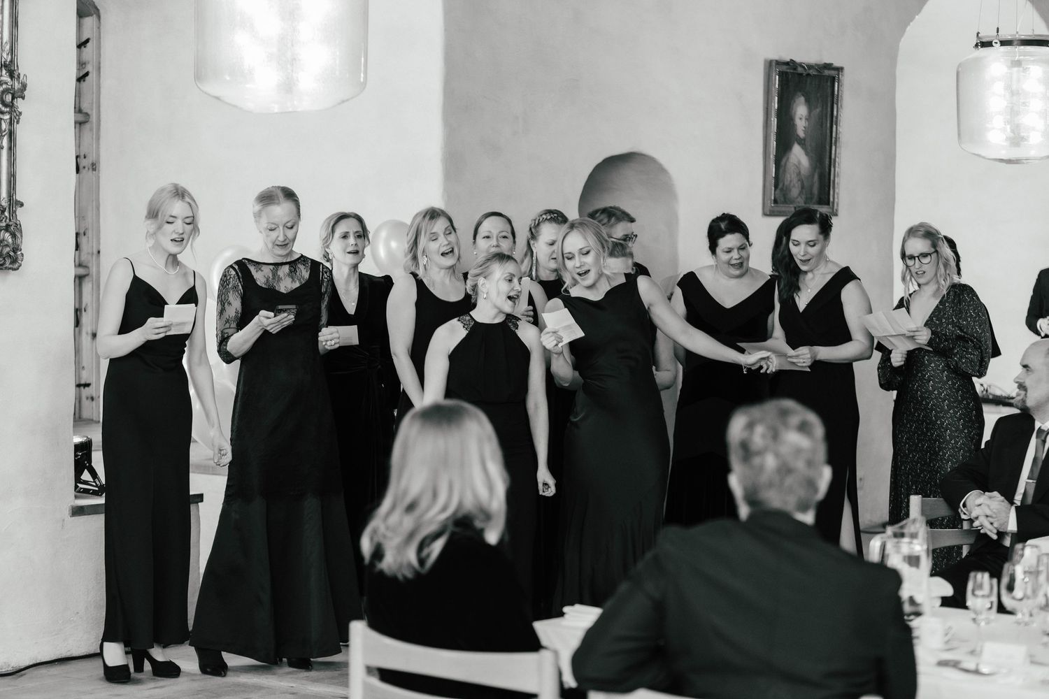 Group of women in elegant black dresses performing or singing at a formal dinner event.