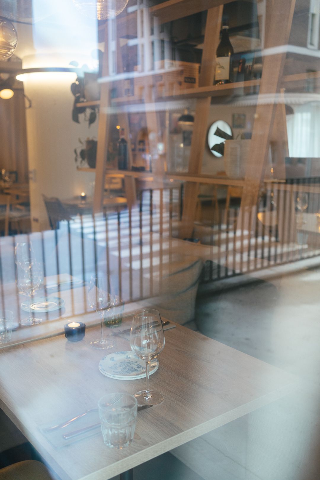 View through restaurant window showing elegant table setting with wine glasses and candle on wooden table.