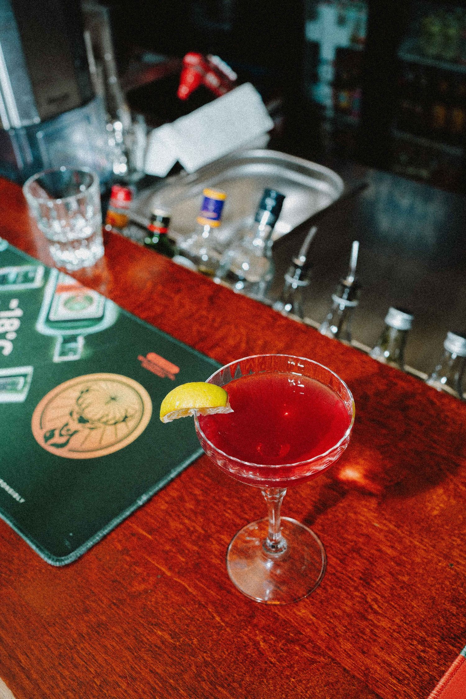 Bright red cocktail with lemon garnish in martini glass on wooden bar counter with Jagermeister mat.