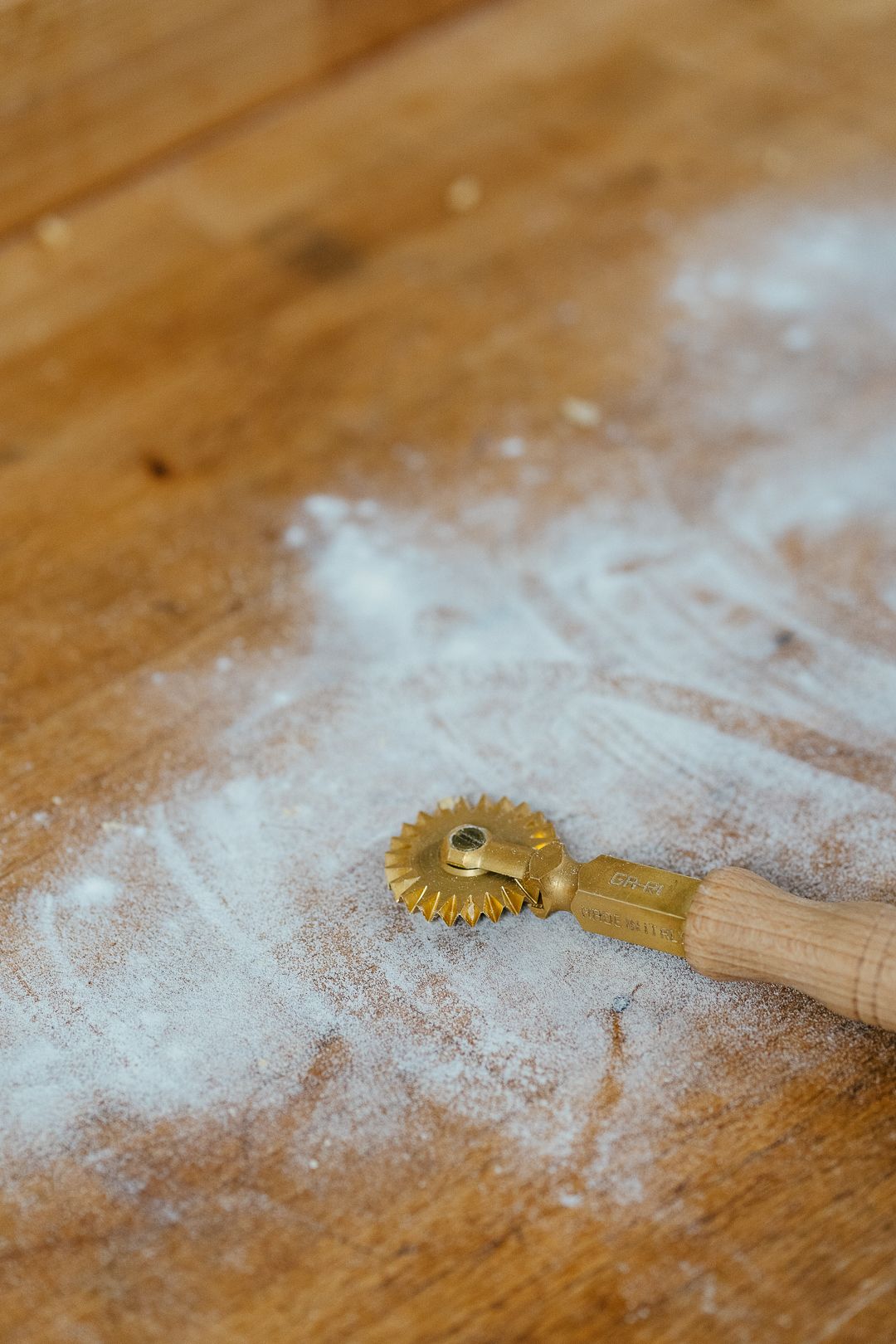 Brass pasta cutting wheel tool dusted with flour on a rustic wooden work surface.