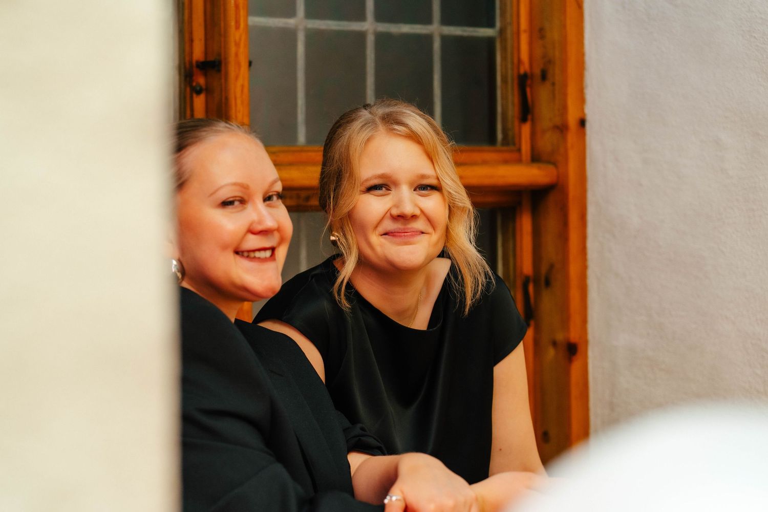 Two smiling women in black attire seated together at a formal event near a wooden window.