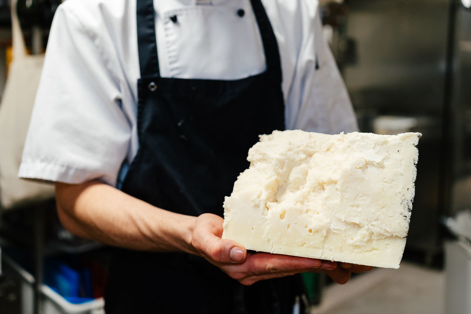 Chef in white uniform and black apron holding a large block of fresh white cheese in kitchen.