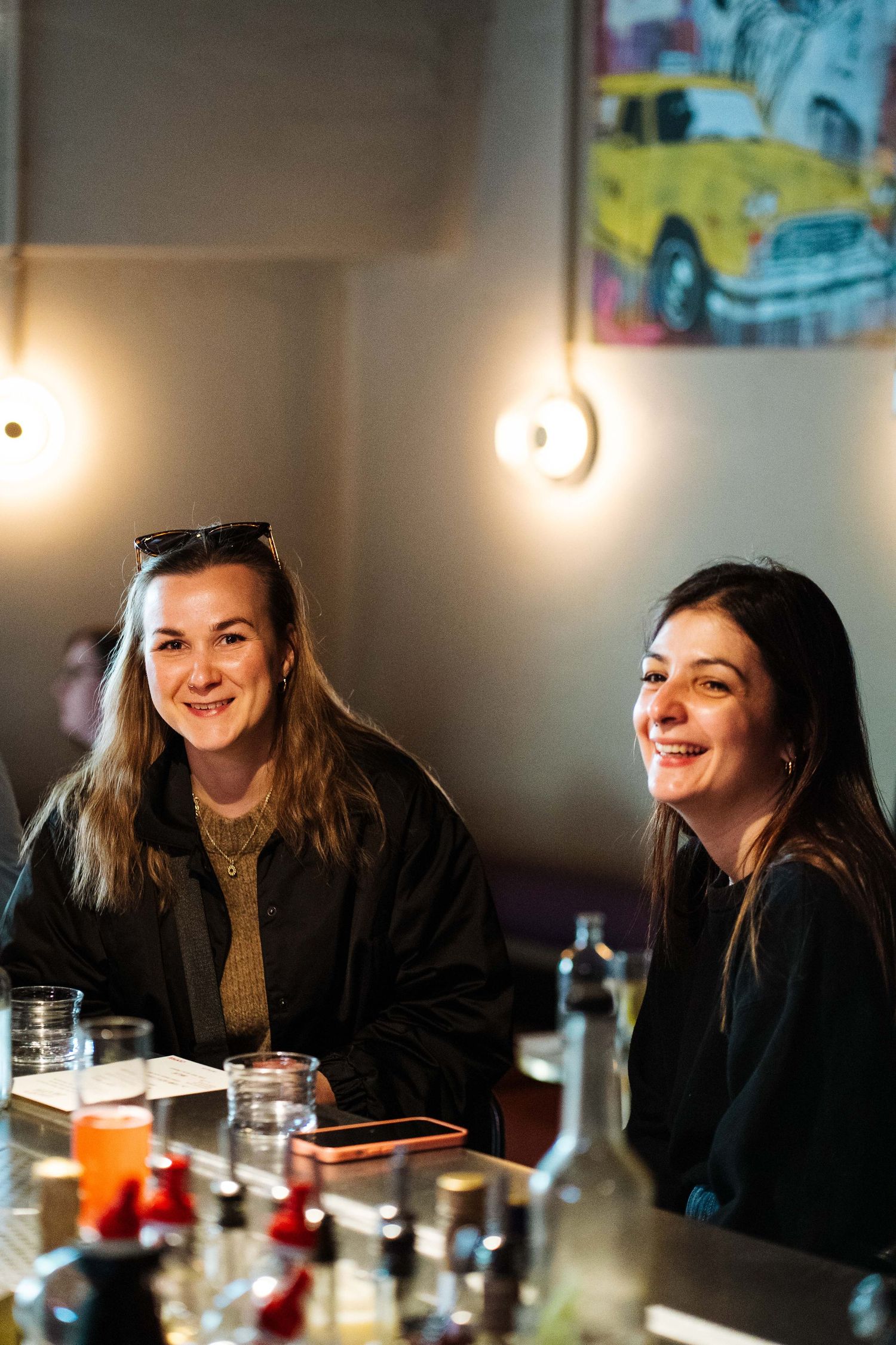 Two smiling women sitting at a bar counter with drinks and bottles in warm ambient lighting.