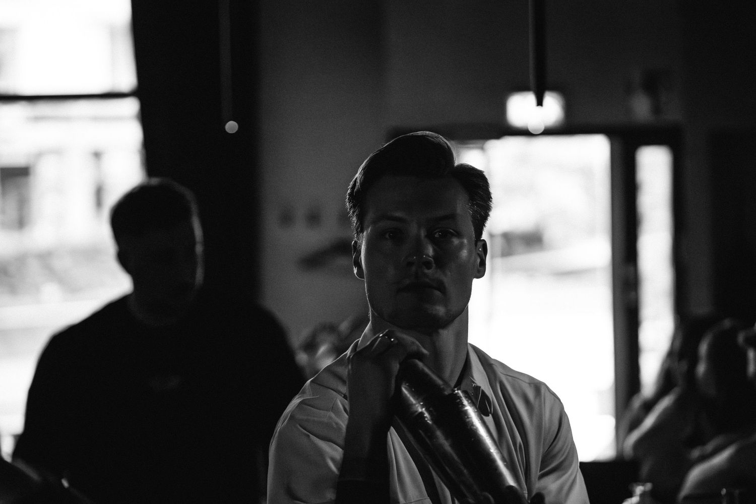 Black and white portrait of a male bartender holding a bottle in a dimly lit bar.