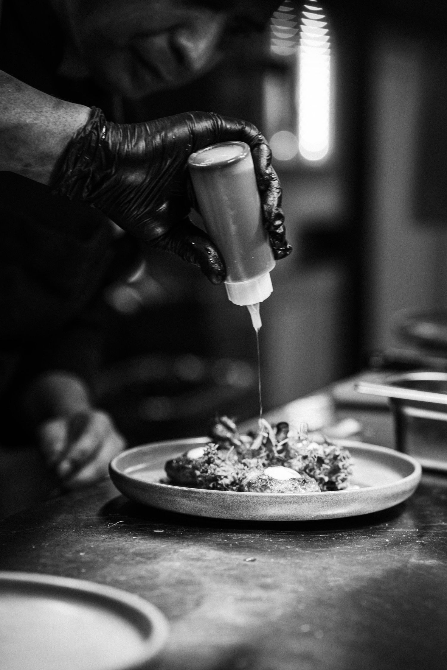 Black and white photo of chef in gloves drizzling sauce over a gourmet dish on a plate.
