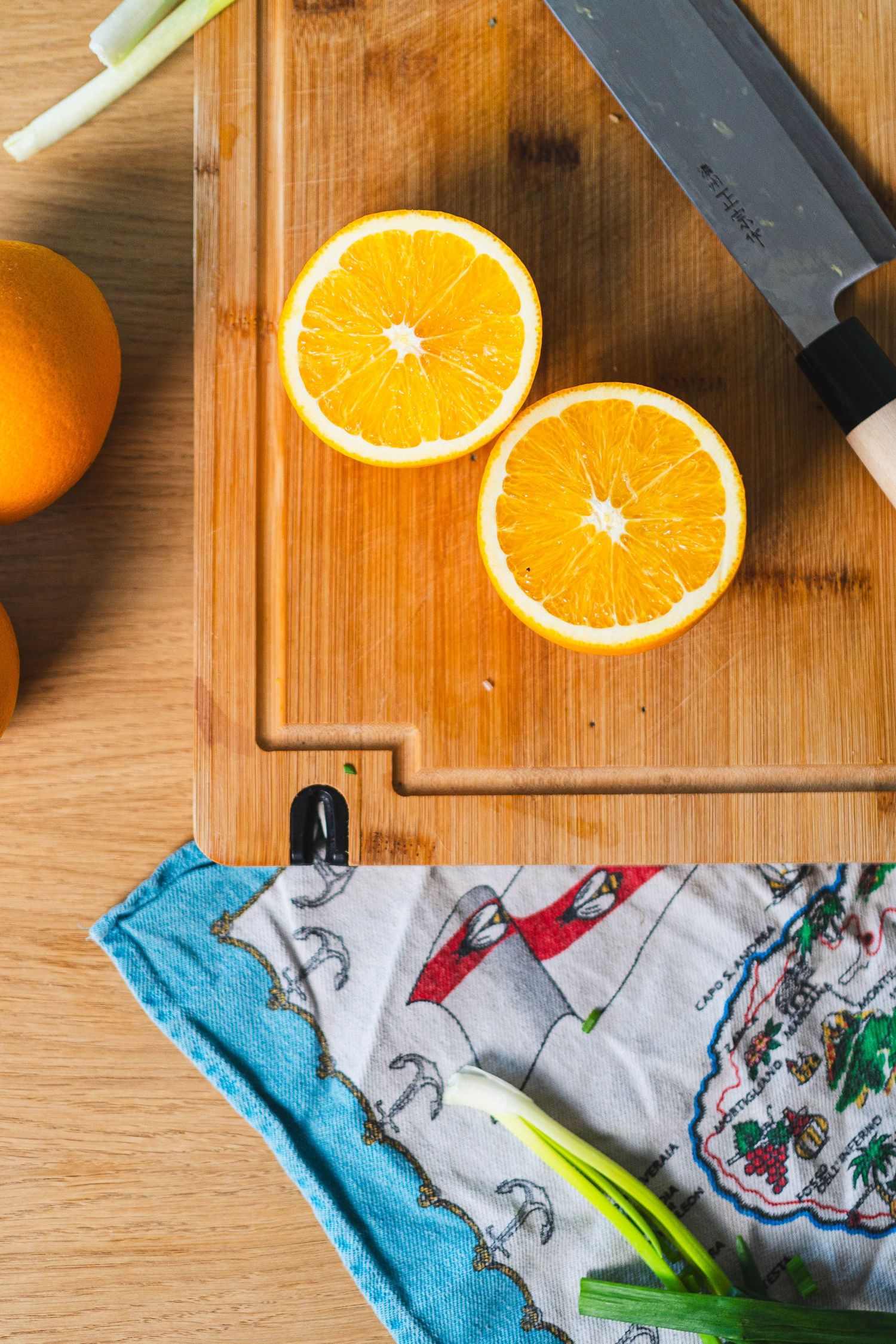 Halved fresh orange on wooden cutting board with knife, green onions and kitchen towel.