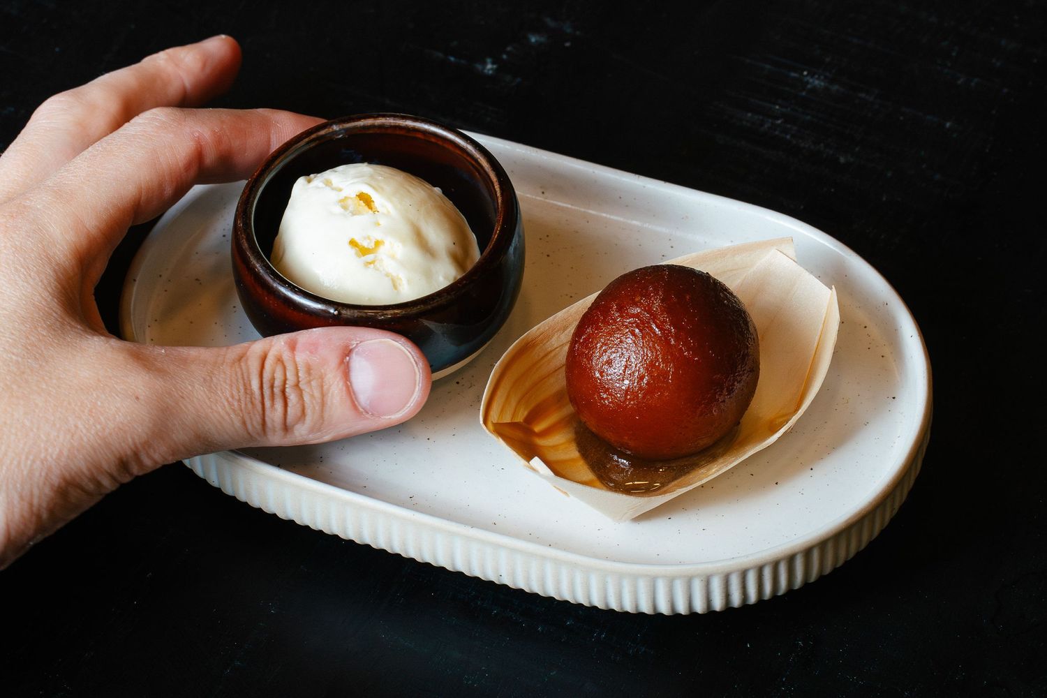 Hand holding a small dessert tray with a gulab jamun and a cup of whipped cream.