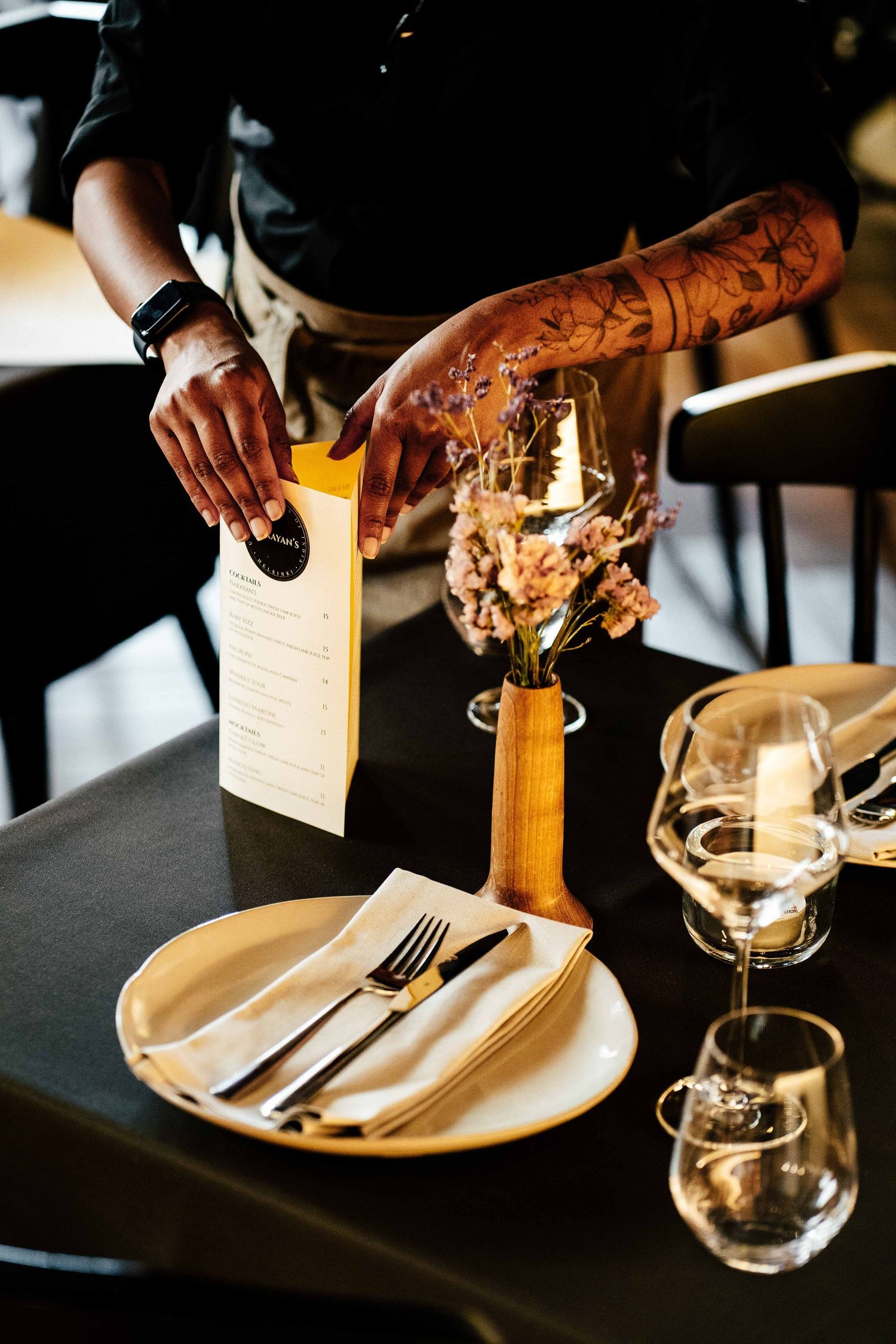 Tattooed server placing a yellow menu on an elegantly set restaurant table with candles.
