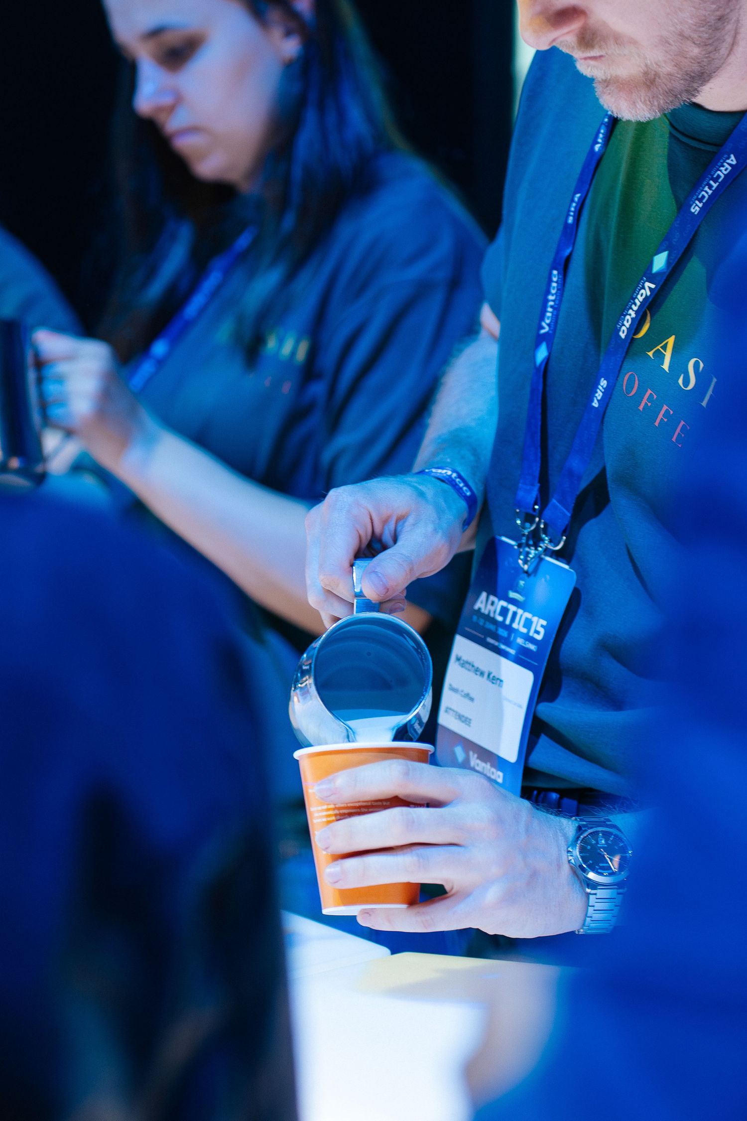 Man pouring liquid into orange cup at a coffee event while wearing Arctic conference lanyard badge.