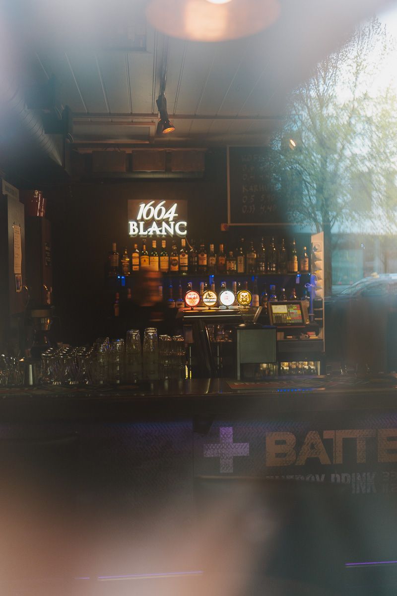 Bar interior viewed through window with 1664 Blanc illuminated sign and bottles on display.