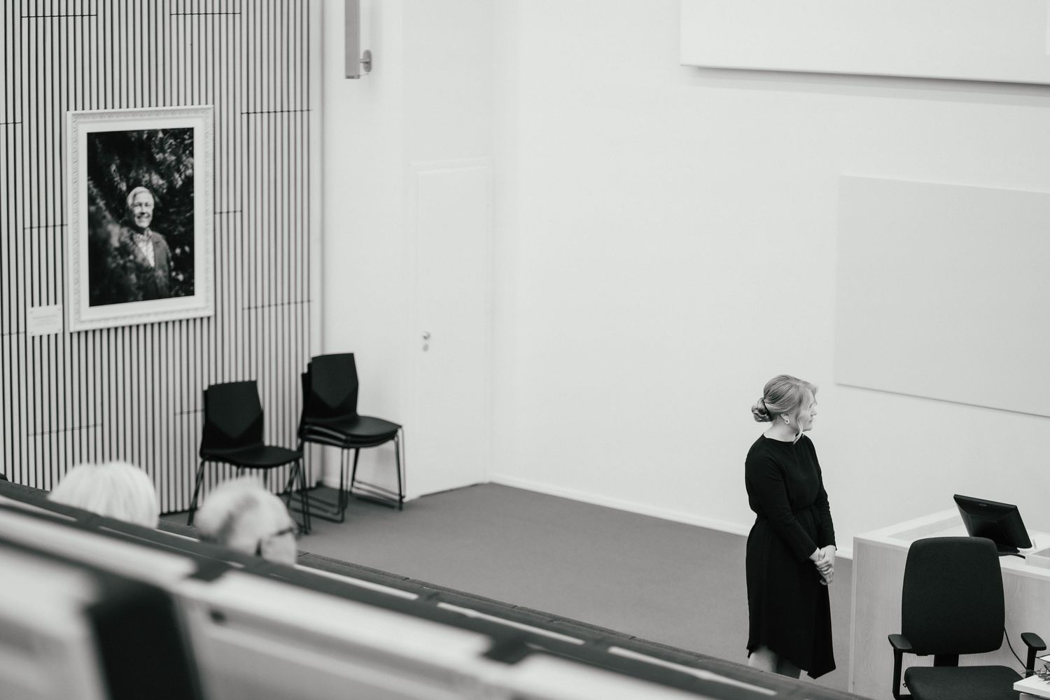 Person in black attire standing in minimalist white gallery space with black and white portrait photograph on wall.