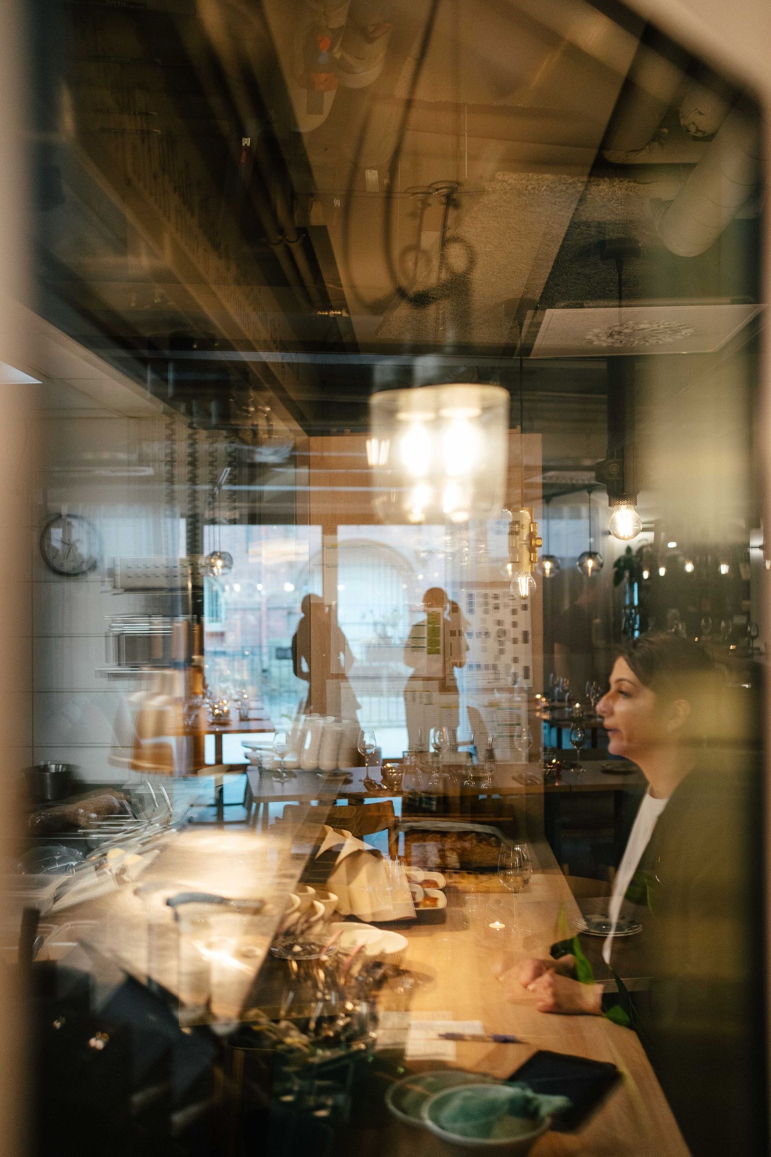 Woman dining in warmly lit restaurant, viewed through glass with reflections of interior decor.