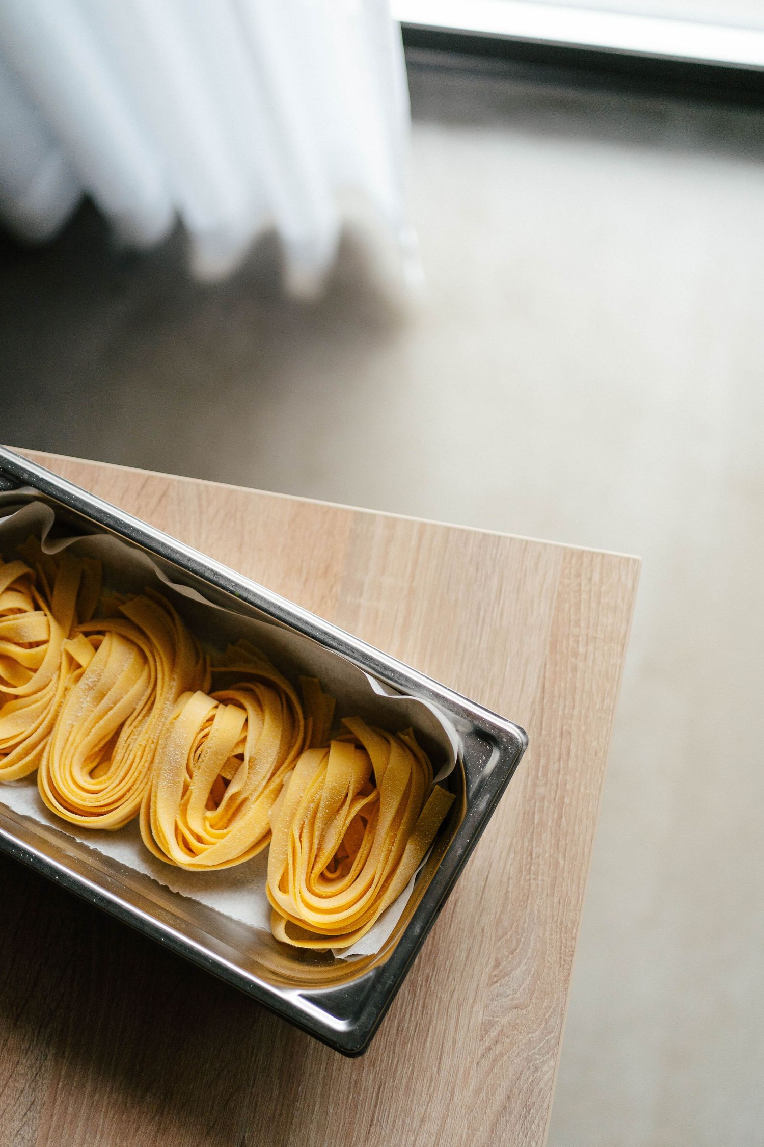 Nests of fresh yellow tagliatelle pasta arranged in a stainless steel gastronorm container on a wooden surface.