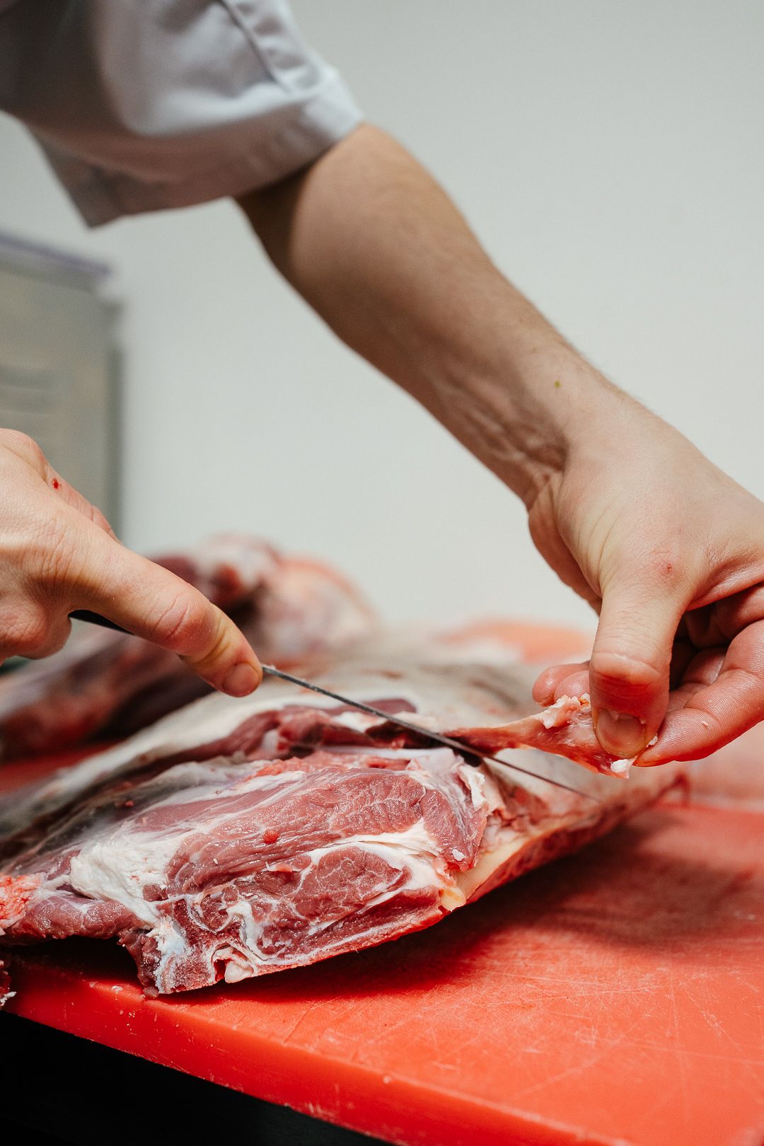 Butcher using a knife to trim and prepare a large raw cut of beef on a red cutting board.