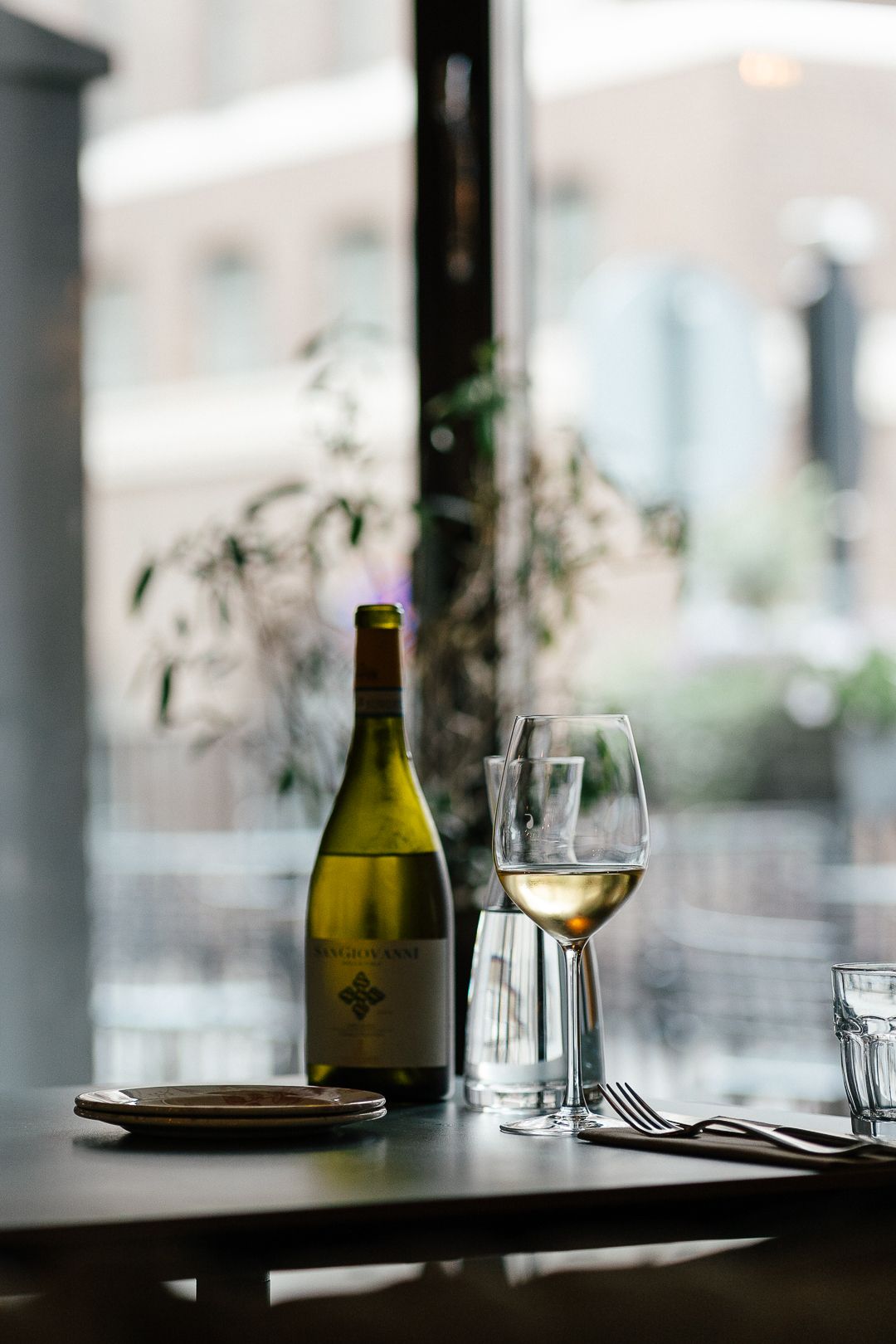 White wine bottle and glass on a restaurant table by a window with city views.