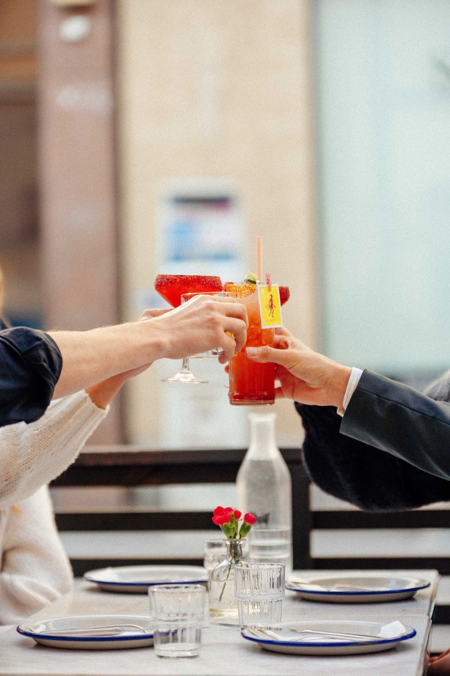 Three hands clinking colorful cocktail glasses in a toast at an outdoor restaurant table.