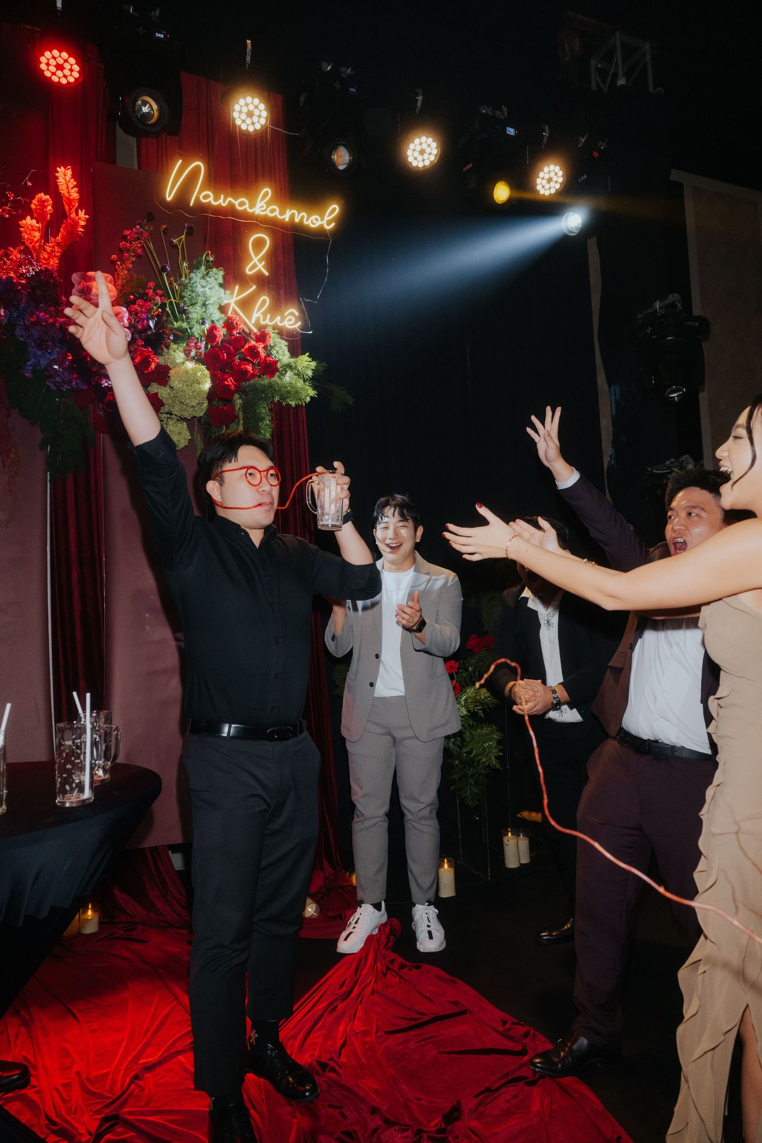 Guests celebrate at a wedding reception with neon sign reading Navakamel & Khue, raising hands in festive toast.