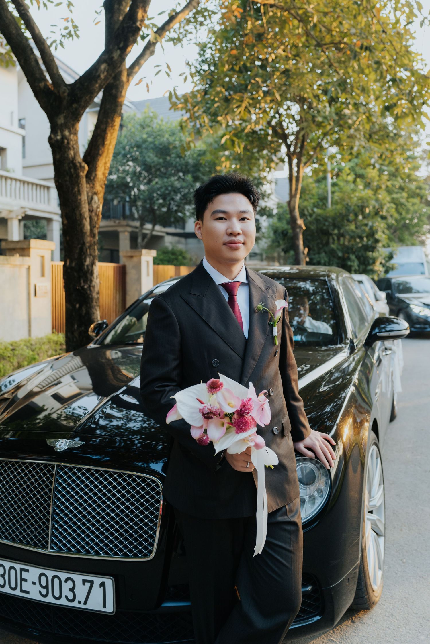Young Asian groom in black suit holding pink bouquet leaning on black Bentley car outdoors.