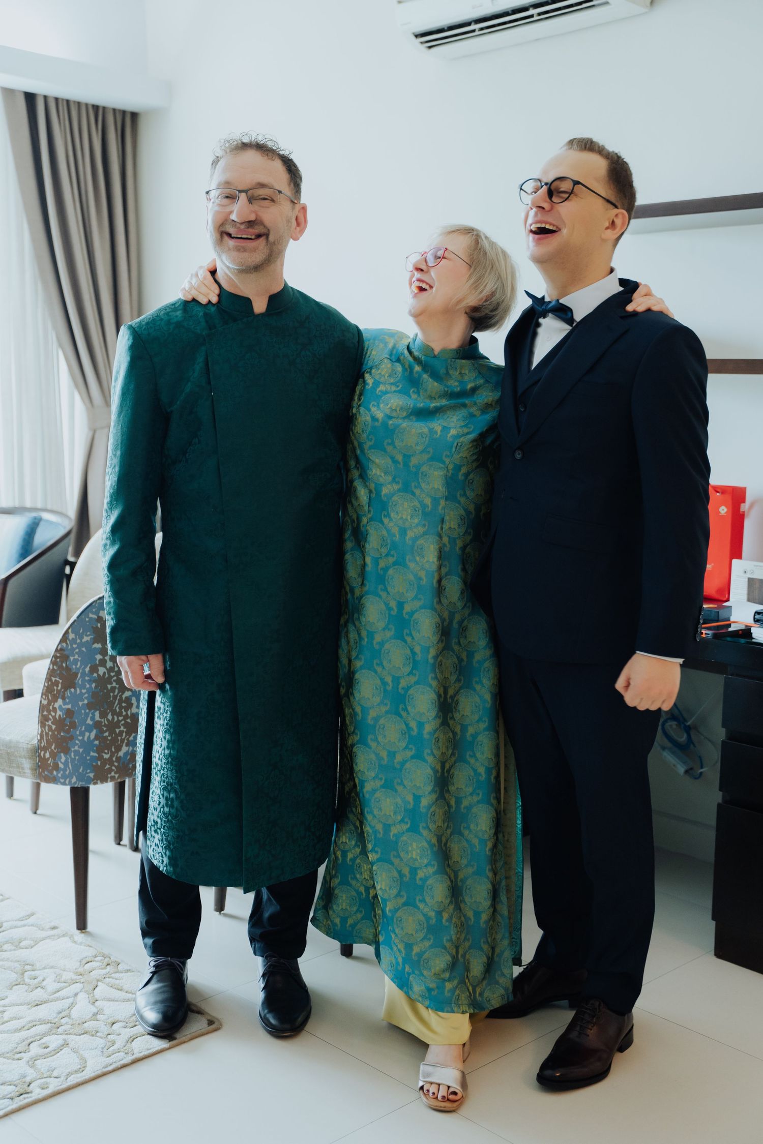 Three people in formal attire smiling together, wearing elegant green traditional outfits and a navy suit at a celebration.