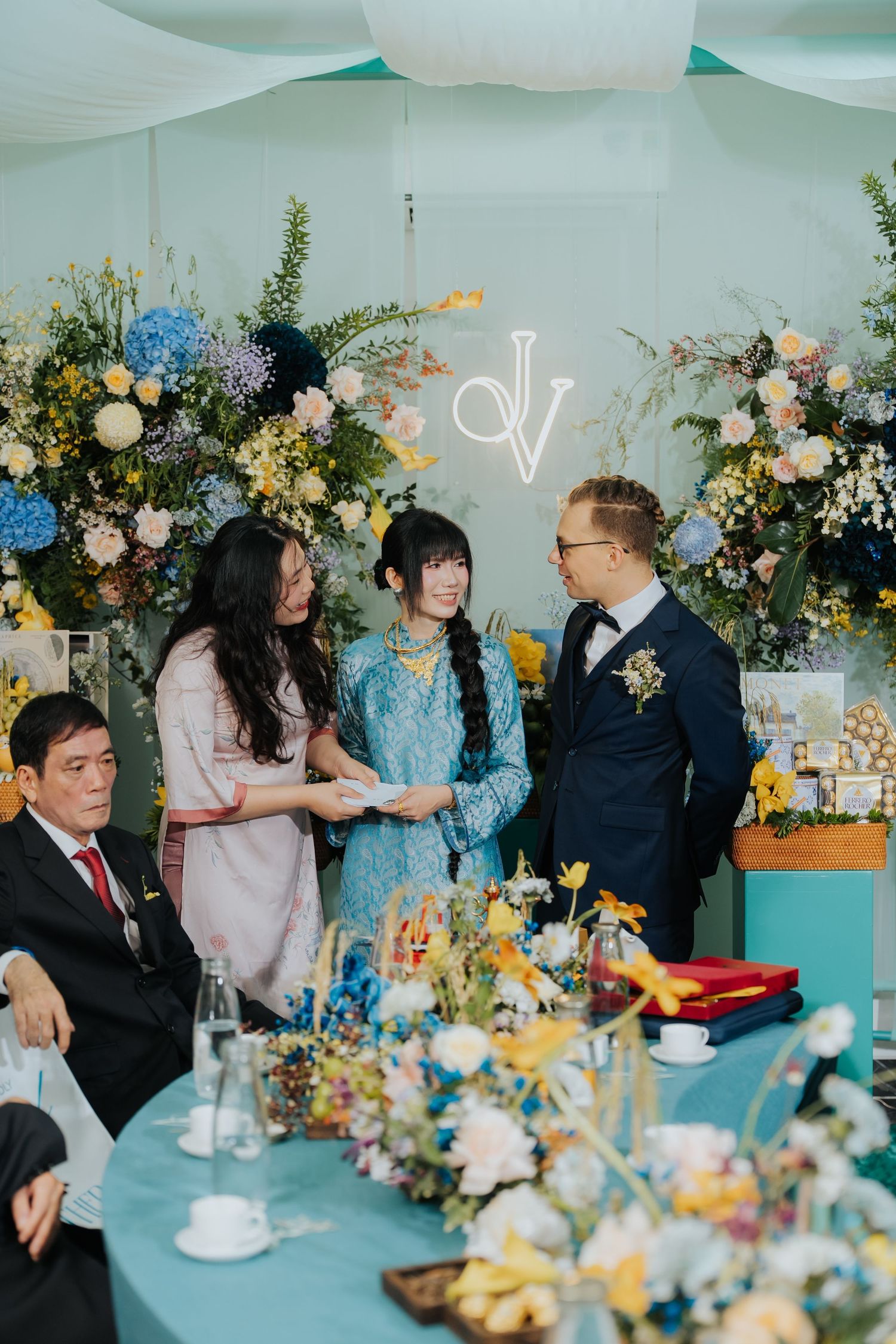 Bride and groom exchange gifts at elegant wedding reception with blue floral backdrop and colorful flower arrangements.