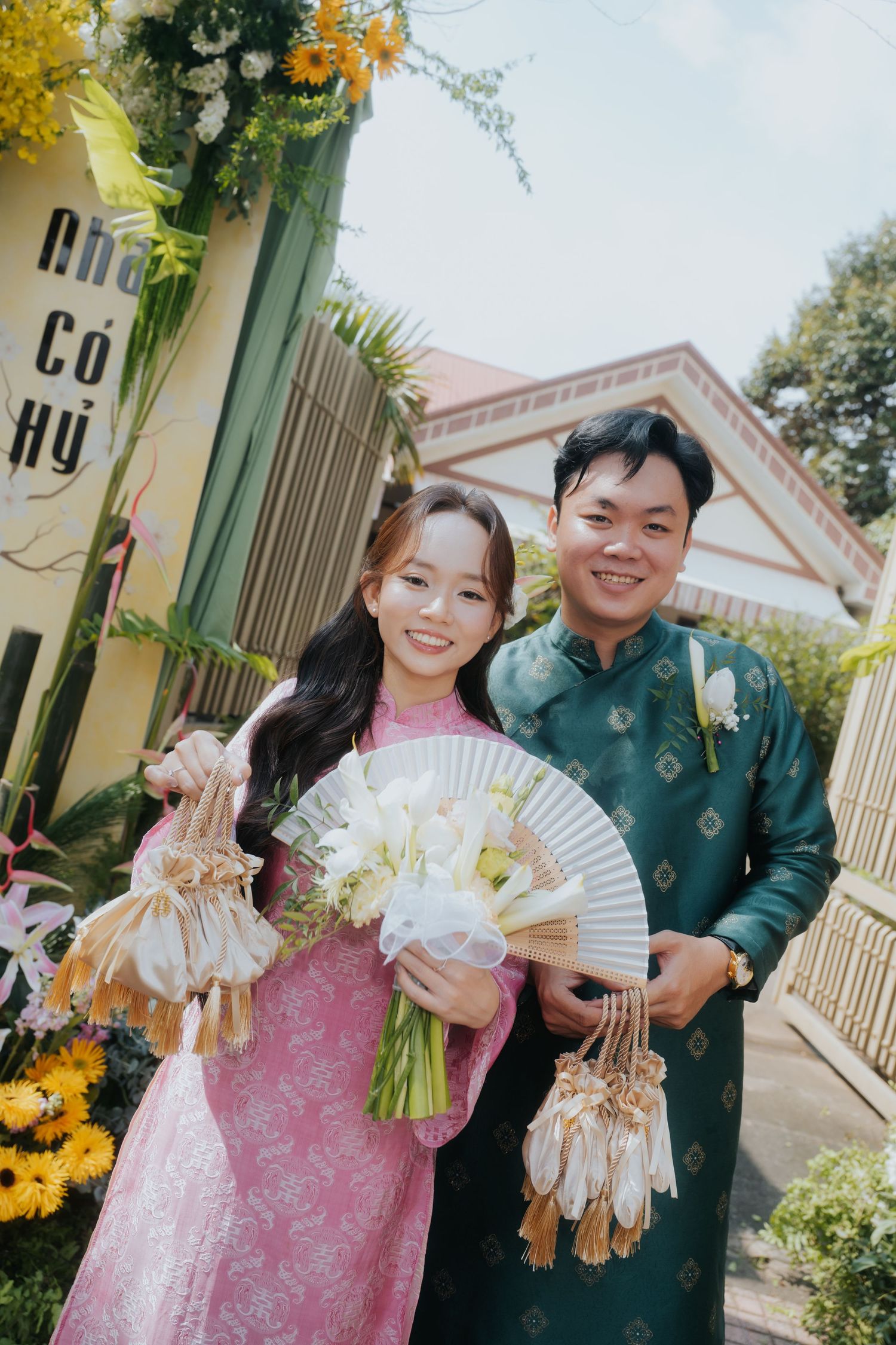 Vietnamese couple in traditional ao dai attire smiling outdoors, holding fans and white tulip bouquet at a wedding ceremony.