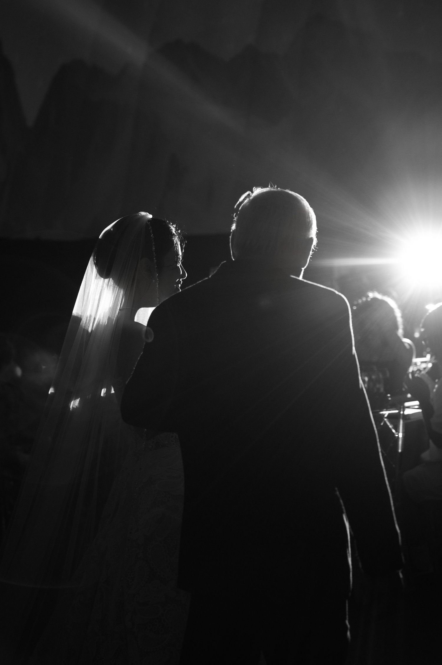 Black and white photo of a bride and older man, likely father, walking toward bright light at a wedding ceremony.