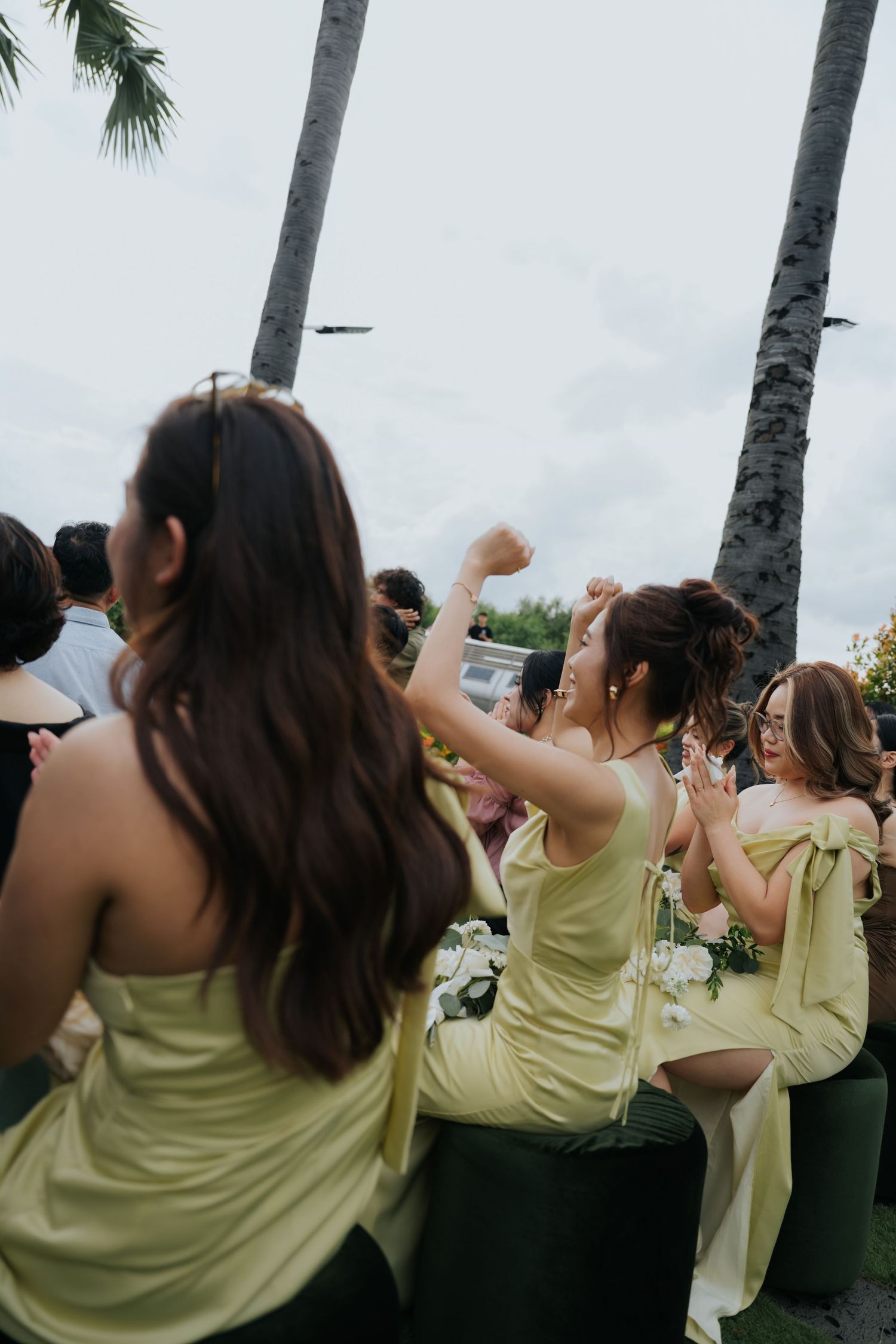 Bridesmaids in matching sage green dresses celebrate outdoors at a tropical wedding with palm trees in the background.