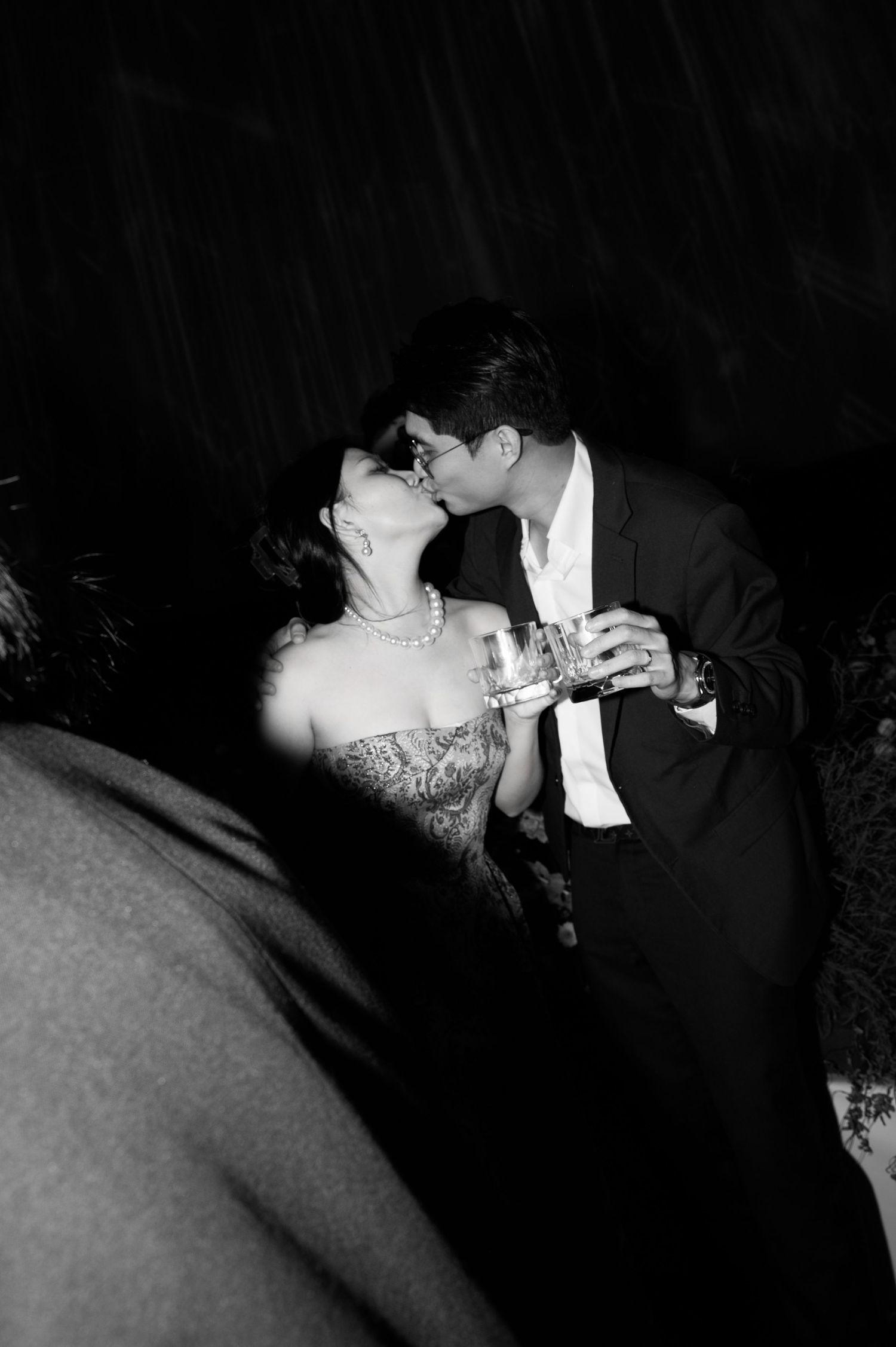 Black and white photo of a couple sharing a romantic kiss at a formal event, holding drinks.