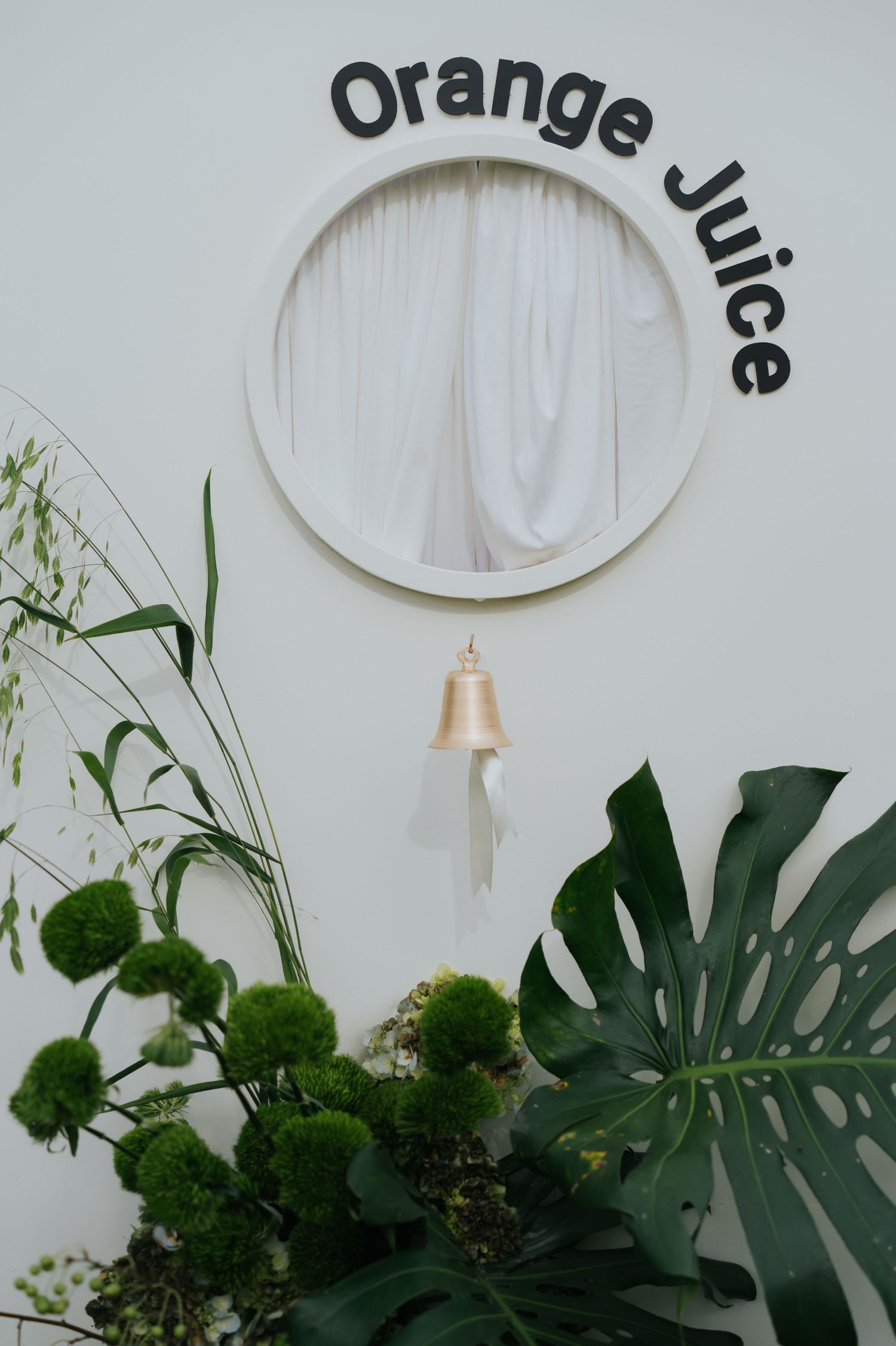 Round window with white curtains, a gold bell, and Orange Juice sign on white wall, surrounded by tropical plants.