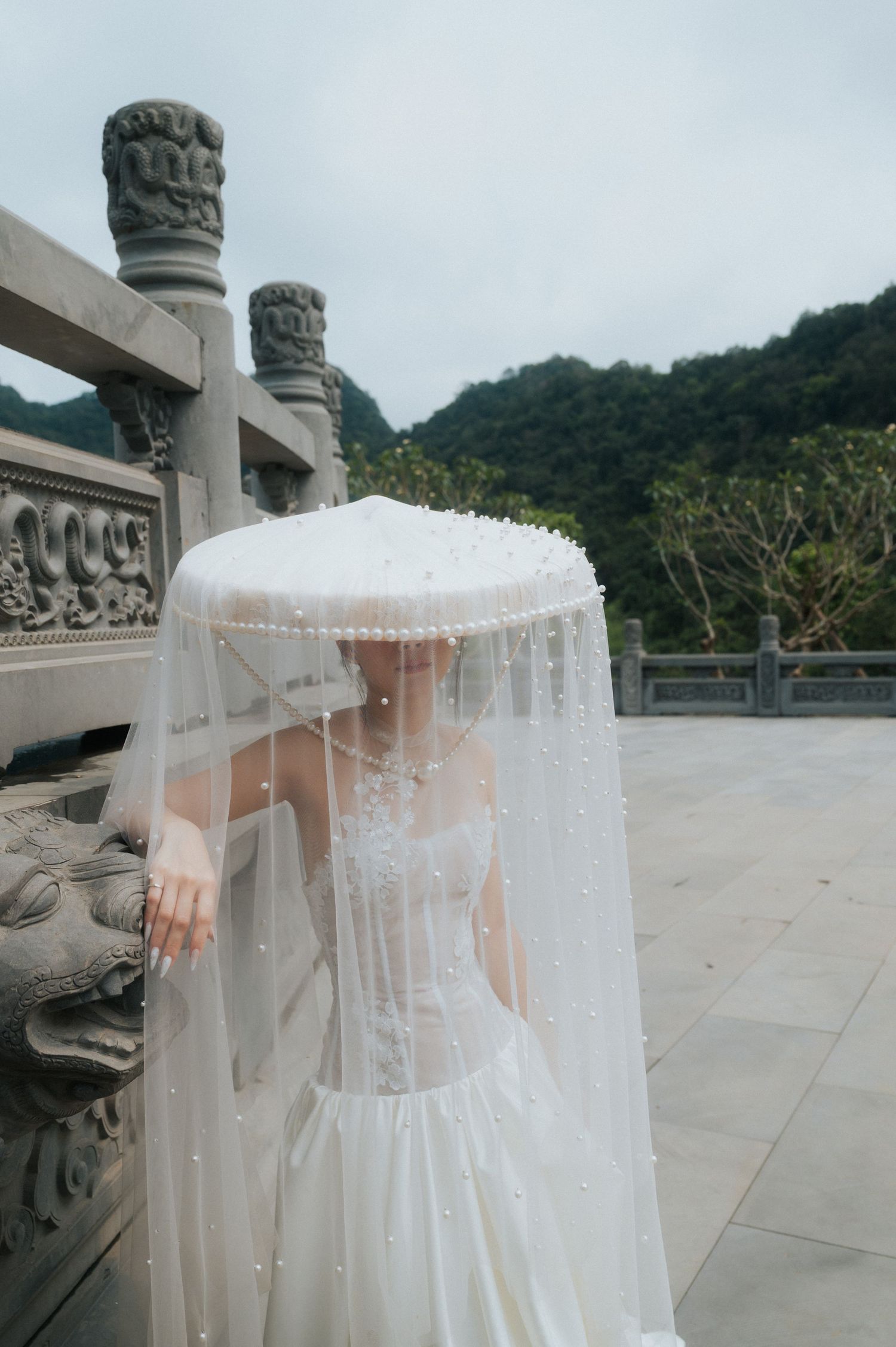 Bride in white wedding dress and pearl-adorned veil at a traditional Asian stone temple with mountain backdrop.
