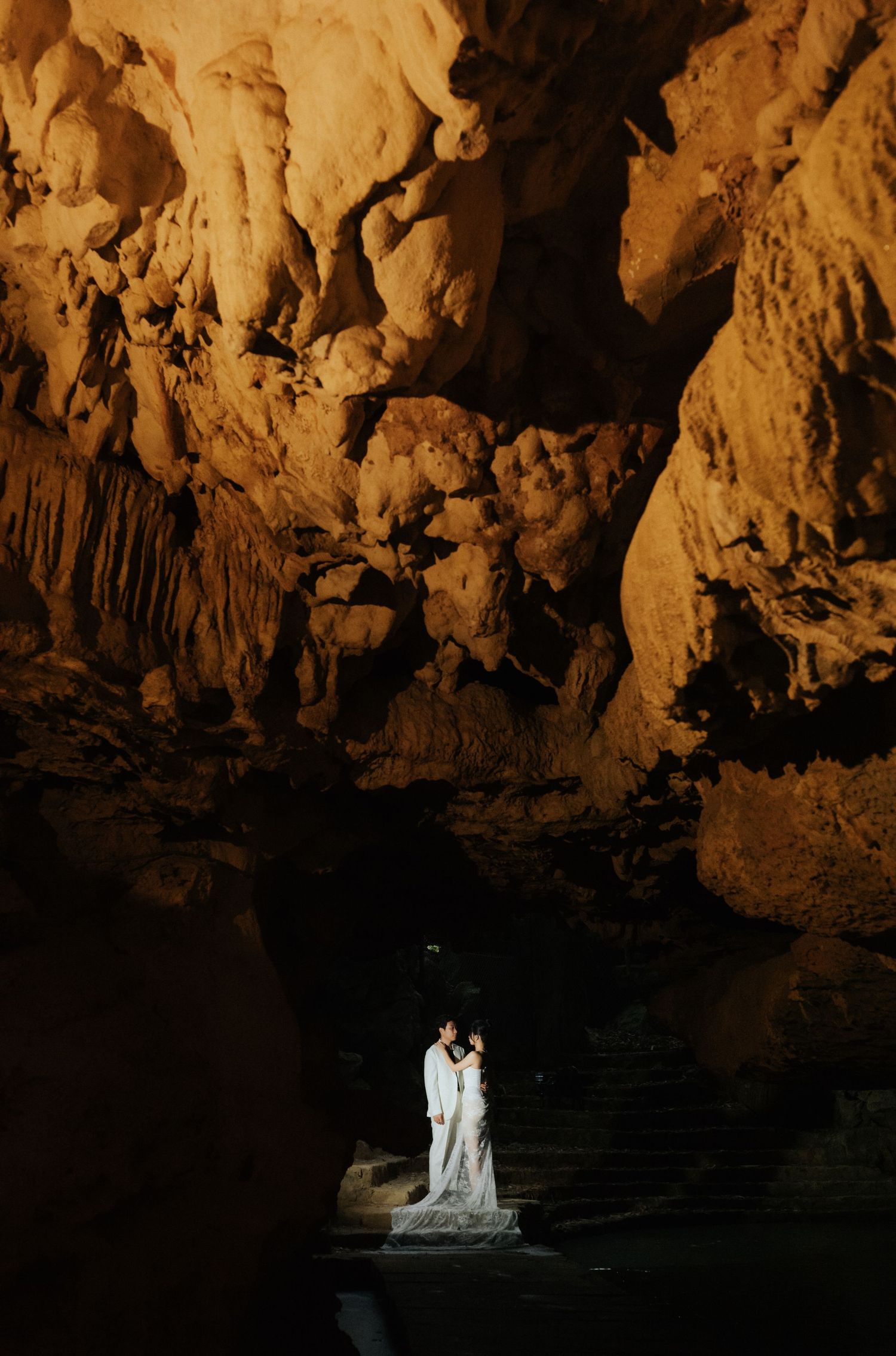 Couple in white wedding attire standing in a dramatic illuminated cave with golden rock formations.