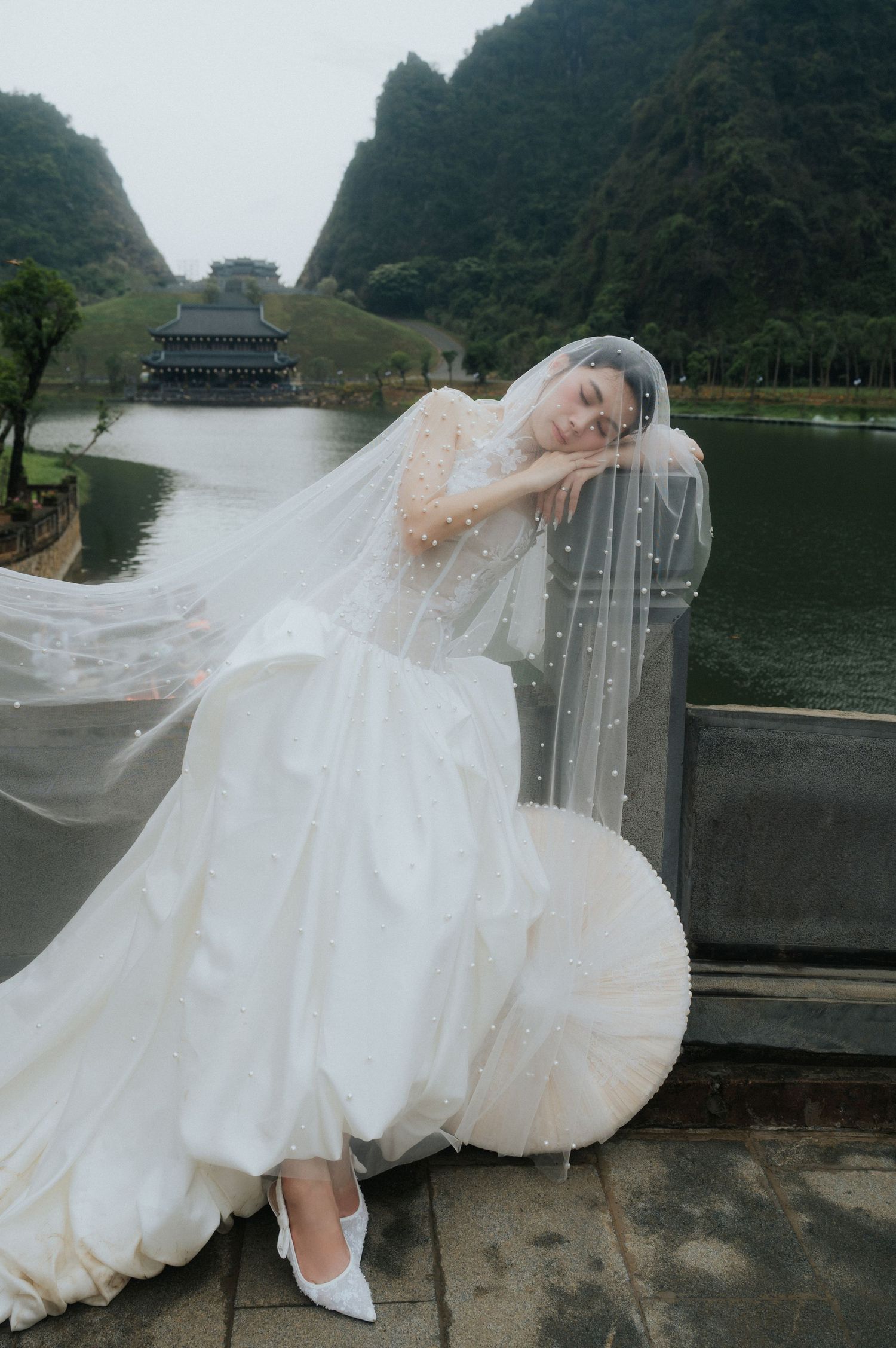 Bride in white wedding gown and pearl veil poses by a lakeside railing with karst mountains in the background.