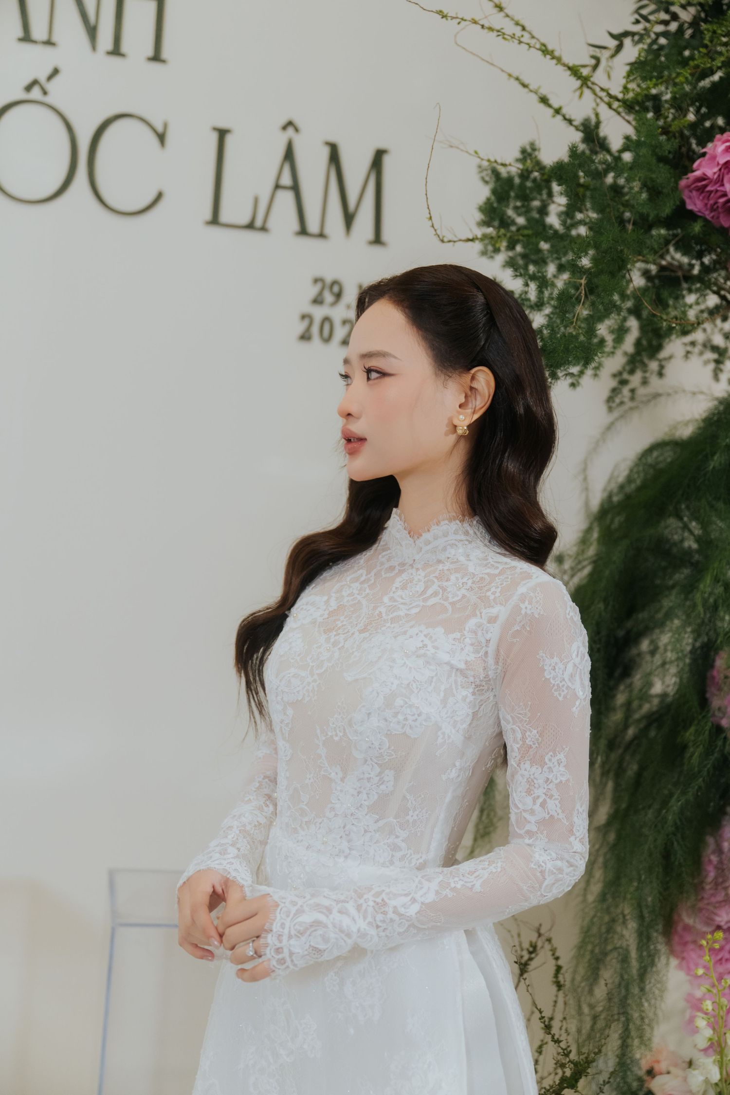 Woman in white lace ao dai gown poses at a Vietnamese wedding ceremony with floral decorations.