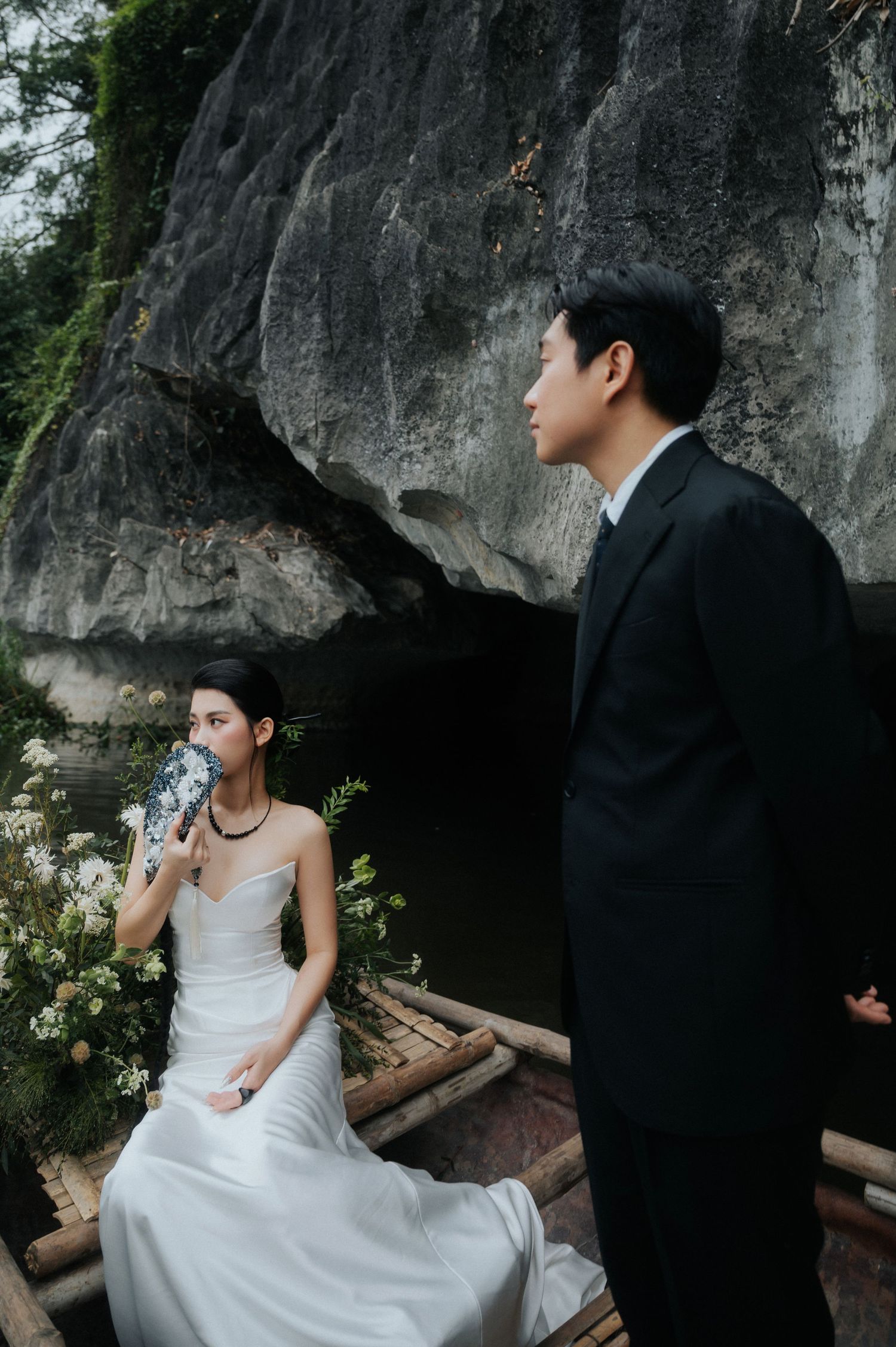A bride in a white gown and groom in a black suit pose near a rocky cave by a bamboo boat with flowers.