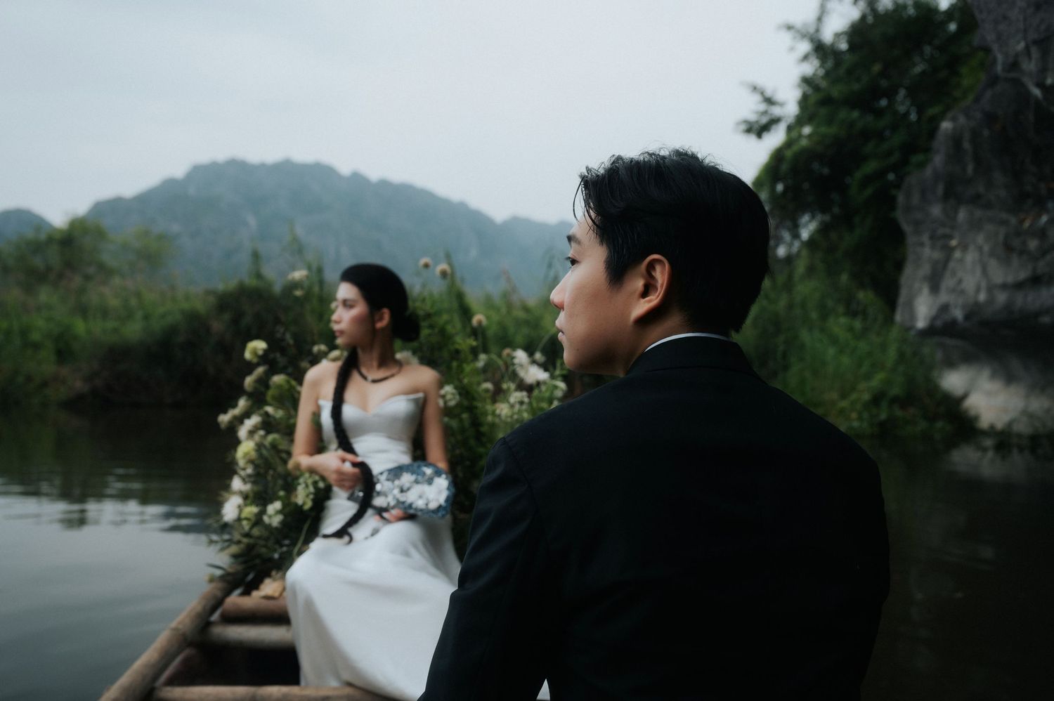 Couple in wedding attire on a wooden boat surrounded by white flowers on a misty river with lush mountains in background.