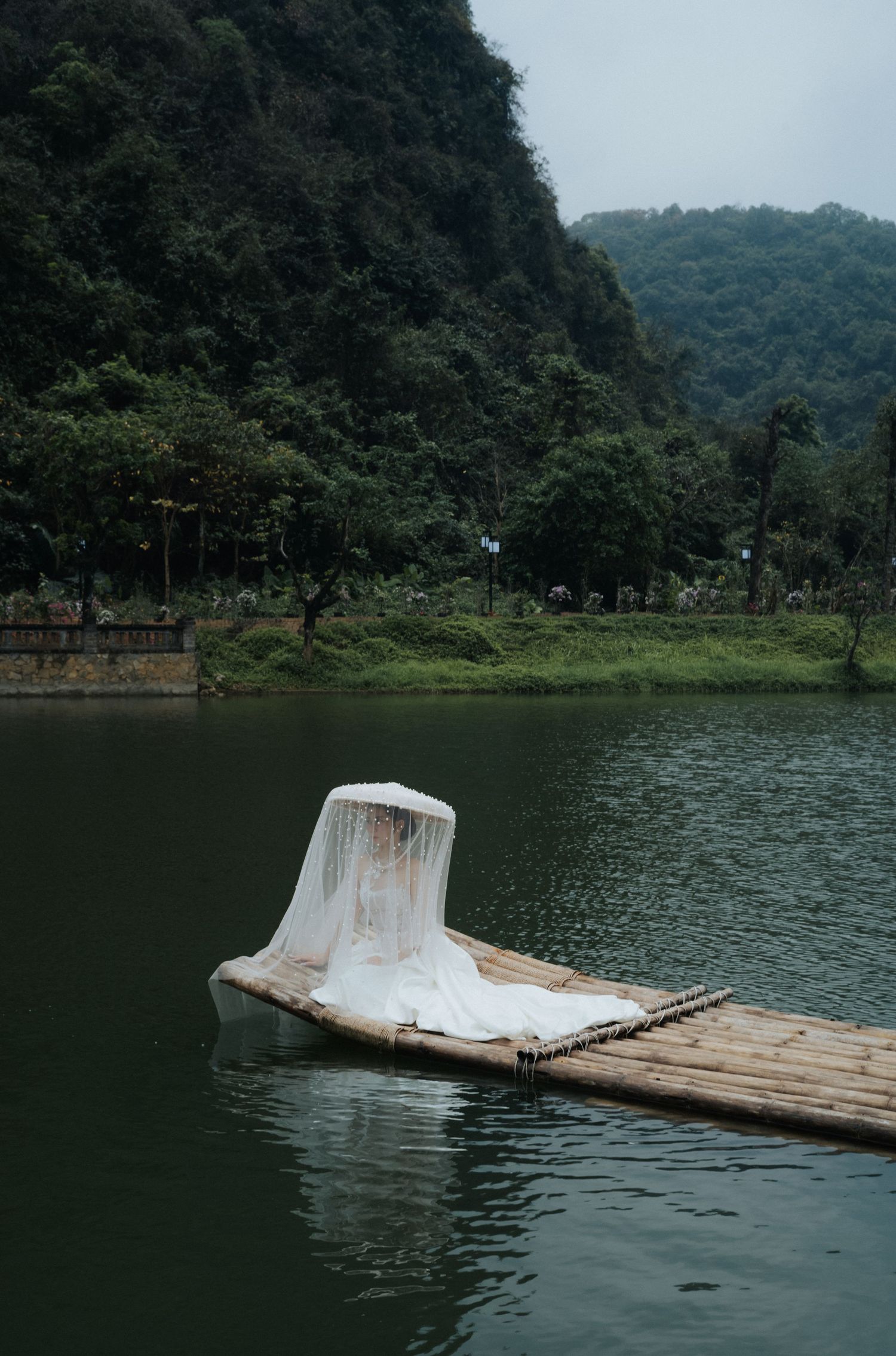 A white draped figure sits on a bamboo raft floating on a calm green lake surrounded by lush forested mountains.