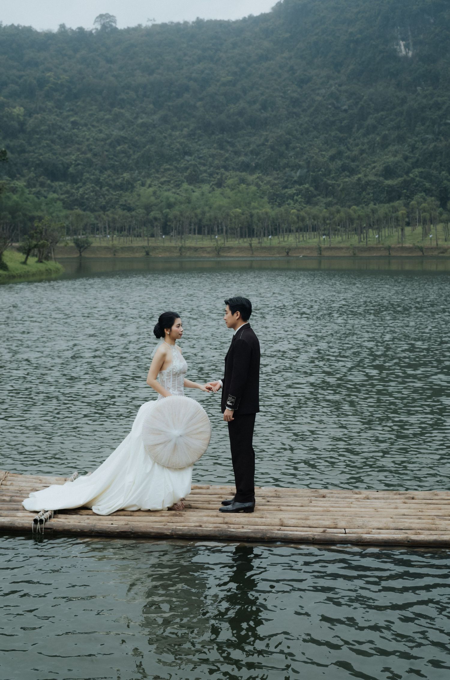 A bride and groom stand facing each other on a wooden dock over a serene lake with lush green mountains behind them.