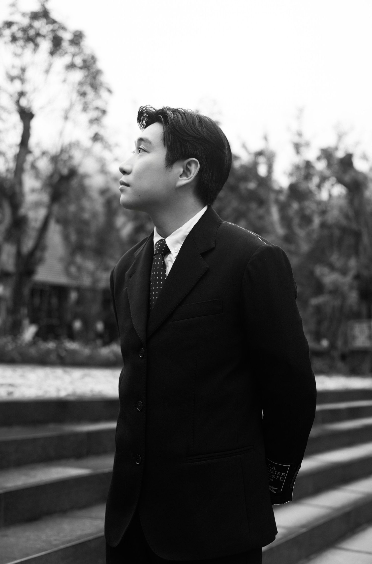 Young Asian man in a black suit and tie gazing upward outdoors in a black and white portrait photo.