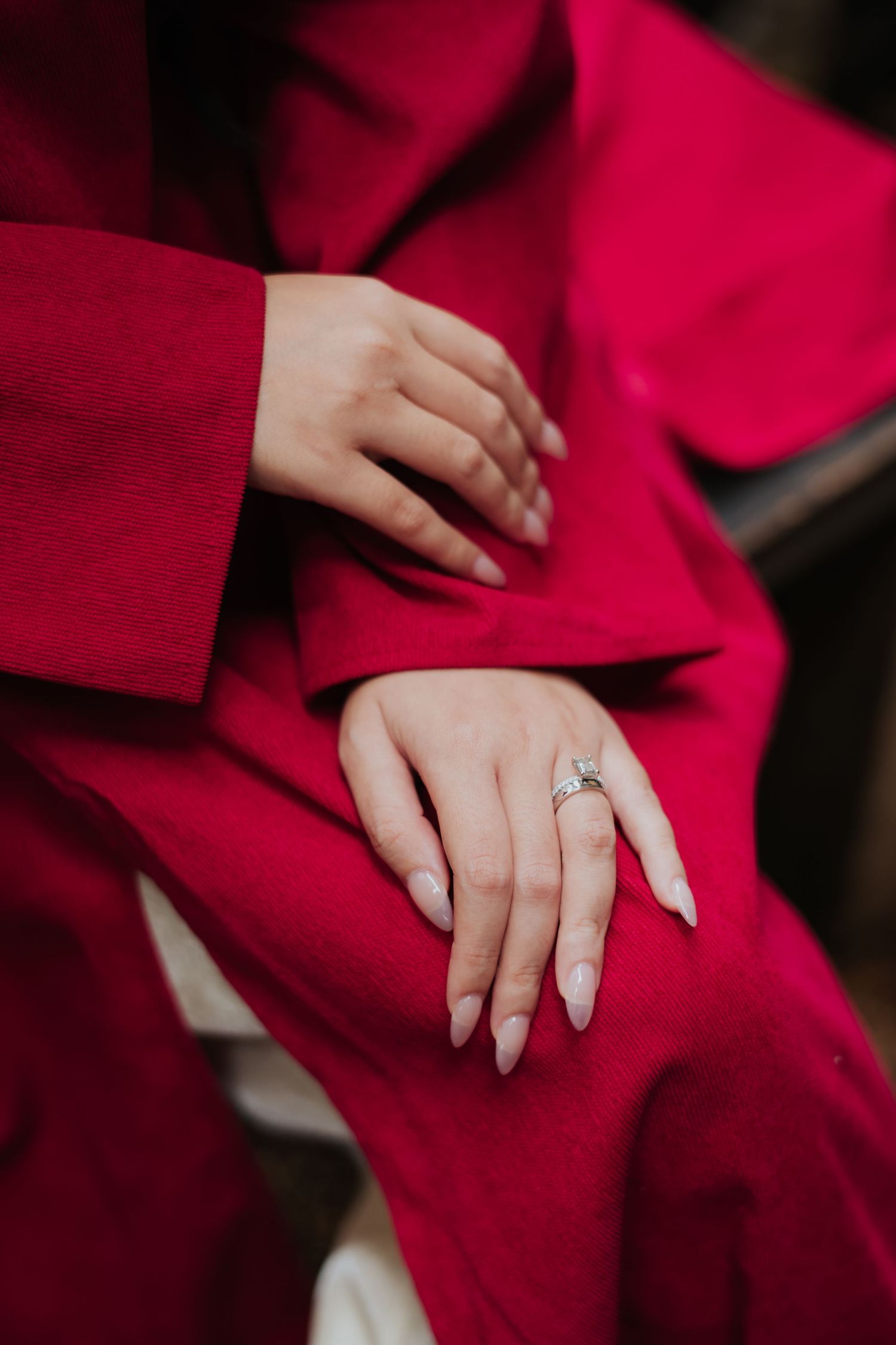 Woman in red suit sitting with manicured hands folded, wearing an elegant silver ring with long nude nails.