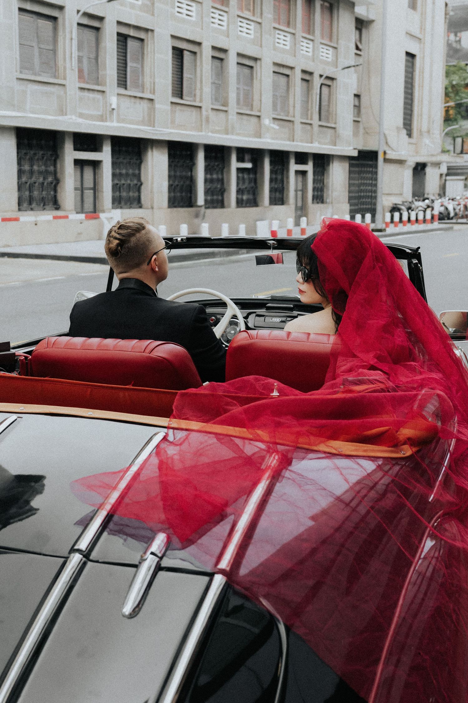 Bride in red veil and groom in black suit ride in a classic red convertible car through city streets.