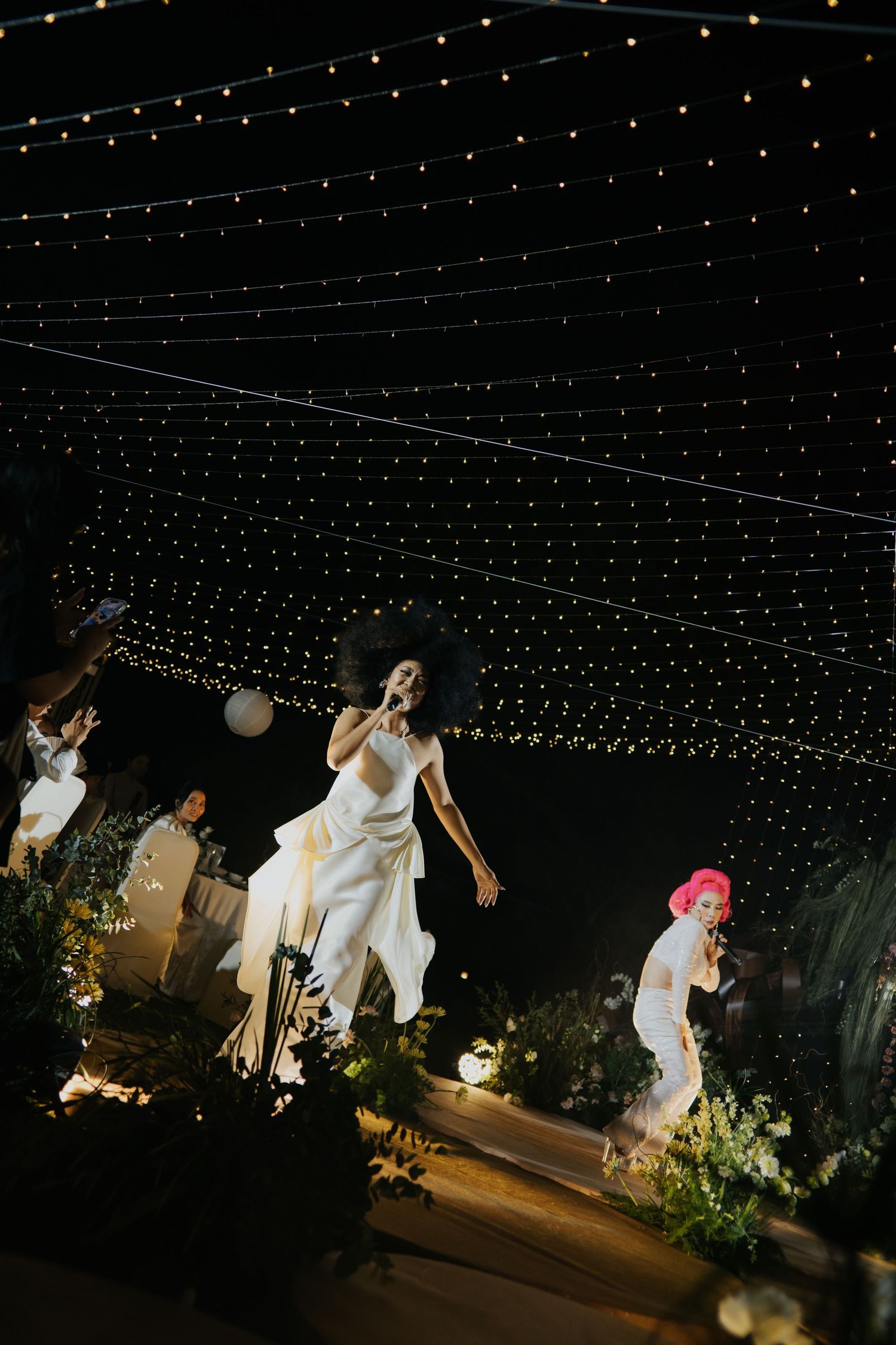Performers in white outfits dance under string lights at an outdoor evening event with lush greenery.